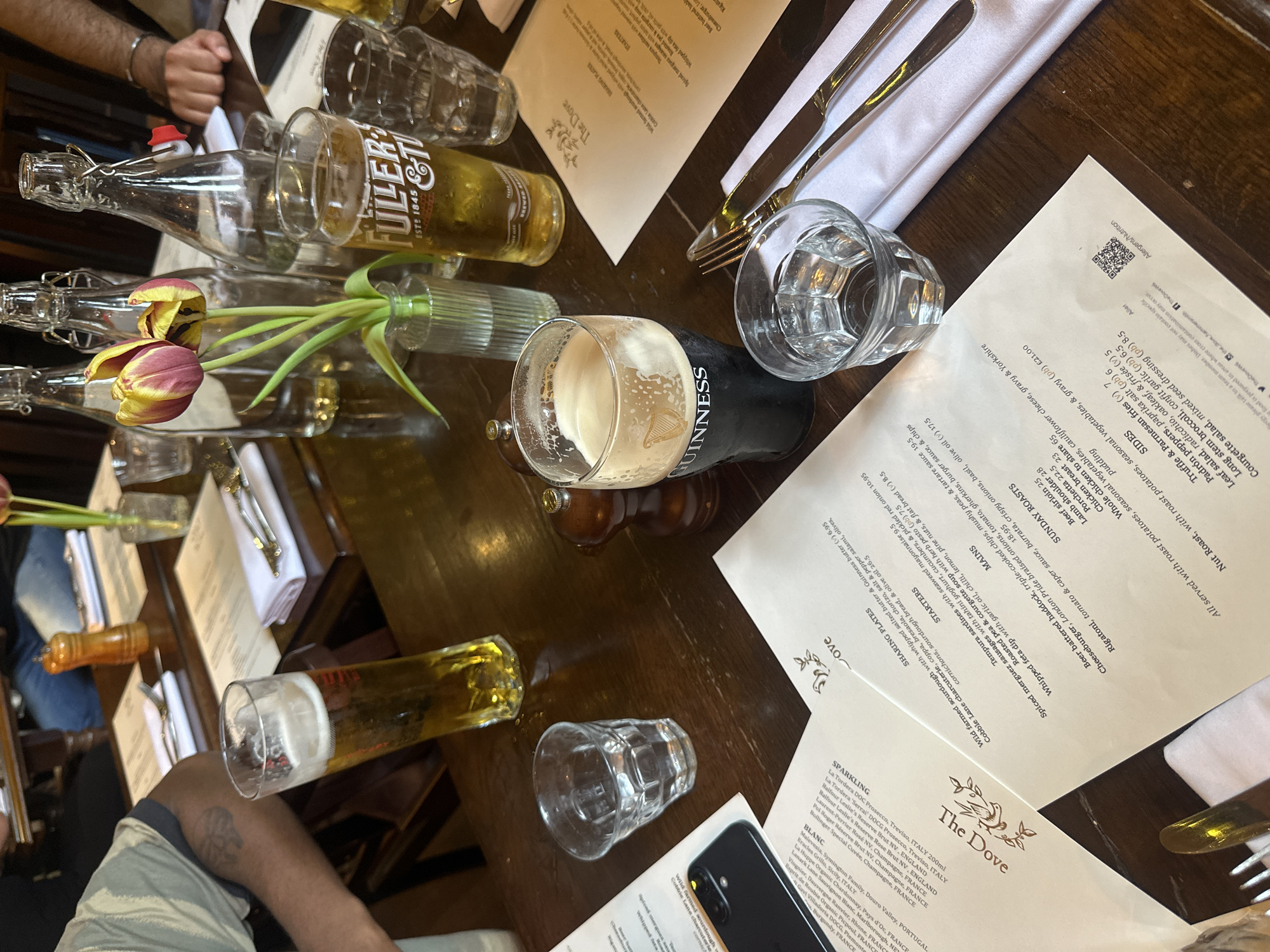 The image shows a table setting at "The Dove" pub in London, United Kingdom. The wooden table is covered with menus, cutlery, a glass of Guinness, a pint of Fuller's beer, and several empty glasses and bottles. Two people's arms are visible at the edges of the frame, suggesting a gathering of at least two people. A small vase with tulips adds a touch of color. The menus list various dishes and drinks, indicating a relaxed dining atmosphere.