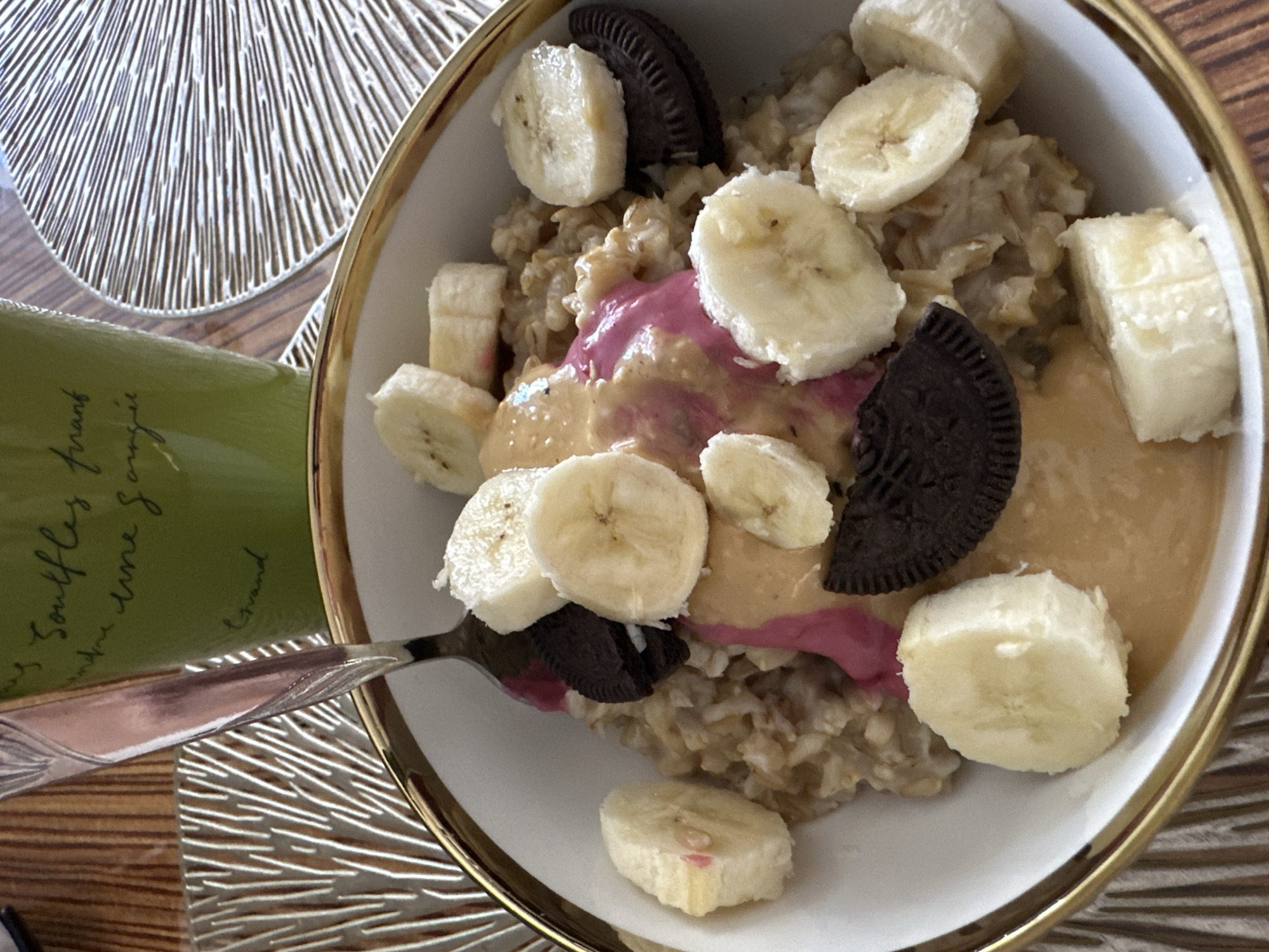 A close-up, overhead shot shows a bowl of oatmeal topped with banana slices, cookies, and a swirl of pink and beige sauces. A fork is partially submerged in the oatmeal, suggesting someone is about to eat it. The bowl has a gold rim and sits on a patterned placemat. In the background, a green mug with handwritten text is visible, also on a wooden surface. The scene appears to be indoors, on a table. The city of Yuzhnouralsk, Russia, is not depicted or suggested by any visual elements in the image.