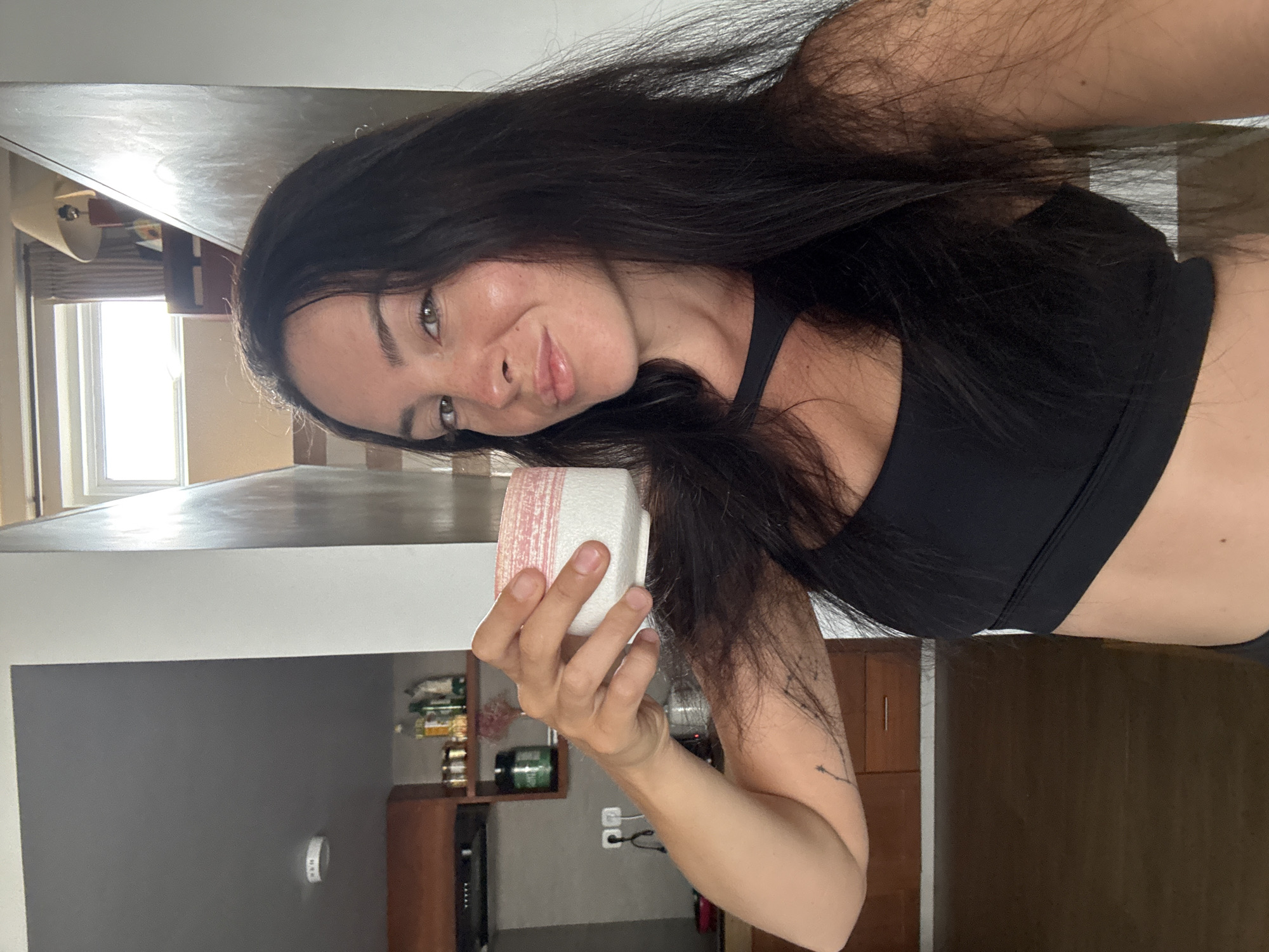 A woman with long, dark hair and light brown eyes is taking a selfie in what appears to be an indoor kitchen area. She is wearing a black sports bra and holding a small, pink and white patterned cup in her right hand, bringing it towards her face. Her left arm is bent, and a tattoo is visible on her bicep. In the background, kitchen cabinets and shelves with various bottles and jars are visible. The lighting suggests it is daytime. The scene is likely located in North Kuta, Indonesia, based on the context provided.