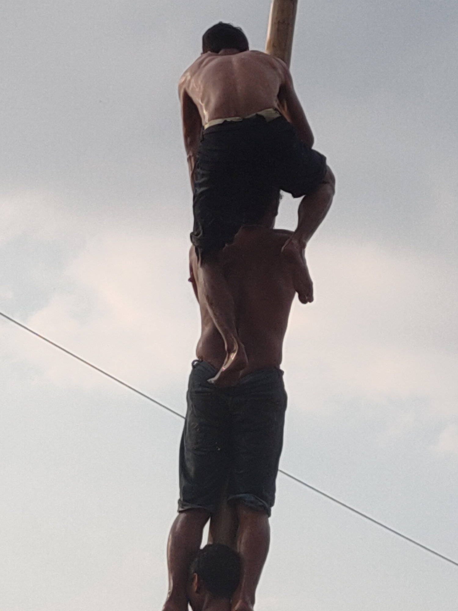 In Kediri City, Indonesia, a group of individuals are engaged in a precarious act of human pyramid formation. The image captures two shirtless men, their bodies glistening with sweat, attempting to climb a tall, wooden pole. The man on top, positioned higher on the pole, wears dark shorts and is seen from the back, his muscular physique evident. He is in the process of hoisting himself further up. Below him, another man, also shirtless and wearing dark shorts, supports the weight of the man above. His legs are clearly visible, gripping the pole. The composition suggests a challenging and potentially dangerous feat, possibly part of a traditional ceremony or performance. The background is a pale, overcast sky with a thin wire stretching diagonally across the frame. The lower portion of the image shows the feet of a third person, hinting at the base of the pyramid.