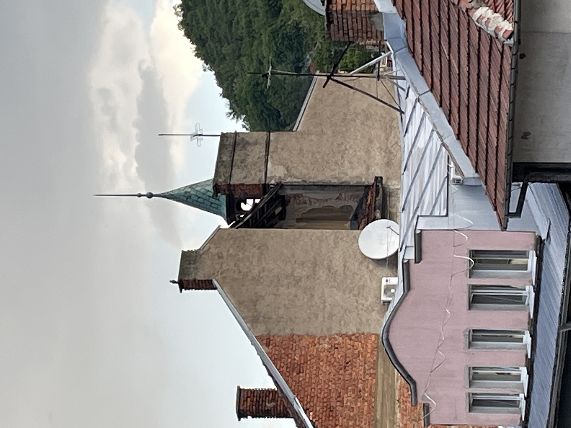 The image displays an overhead view of a cluttered urban rooftop landscape under an overcast sky. In the left-center, a building with a distinctive green, conical spire, topped with a sharp finial, dominates the frame. Below the spire, the structure features an aged facade of light brown stucco with sections of exposed red brick, showing signs of wear or renovation.
To the right, various other buildings contribute to the dense cityscape. One building on the right has a pink/salmon-colored facade with multiple rectangular windows, and a white satellite dish is mounted on its wall, with a white air conditioning unit positioned underneath. Another building displays a broad, light brown stucco wall. A mix of red-tiled roofs and grey corrugated metal roofs are visible across different structures.
Several antennas, including a cross-shaped one and a Yagi-style antenna, protrude from the rooftops, indicating communication infrastructure. Gutters, downspouts, and a wavy white cable are also present. In the upper background, a lush green hillside densely covered with trees provides a natural backdrop against the muted grey and white clouds. The overcast conditions suggest daytime, possibly early morning or late afternoon, with a soft, diffused light. There is no human activity or visible text in the scene.