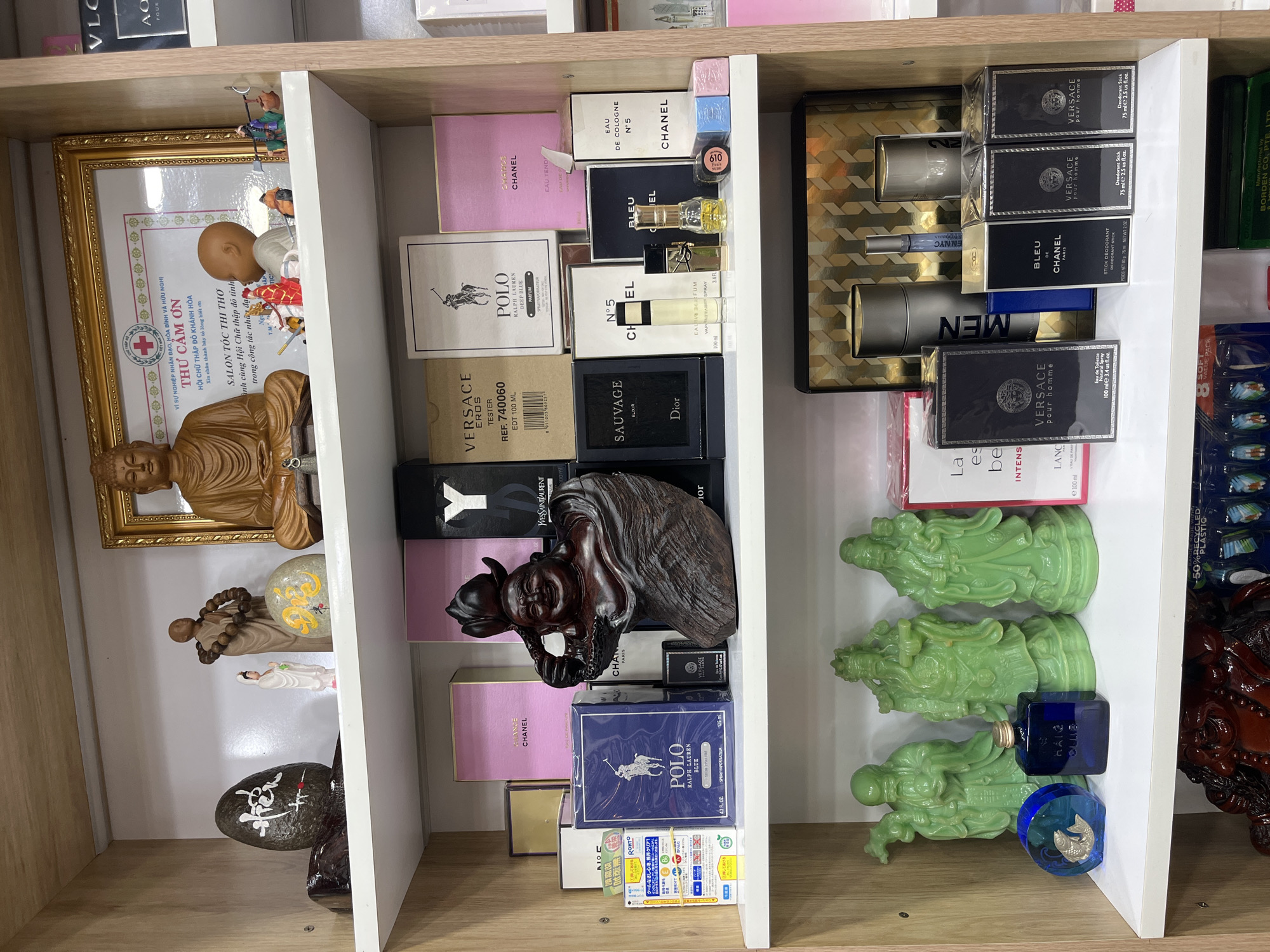 The image shows a retail shelf displaying various perfumes and decorative items. In the top shelf, a framed certificate is prominently displayed, featuring text in Vietnamese and a red cross emblem. Beside it, a wooden statue of Buddha sits, and to its left are smaller figurines of people and animals. Further left, a dark stone with white calligraphy and a light-colored, round stone with yellow calligraphy are visible. The middle shelf is filled with perfume boxes of various brands, including Chanel, Versace, and Polo Ralph Lauren, as well as a wooden carving of a laughing figure. The bottom shelf features several green figurines resembling deities, a blue perfume bottle, and more perfume boxes, some with the Versace logo. The shelves are made of wood. The background is a white wall with some wooden paneling on the right. The items are arranged neatly. The image is taken in Nha Trang Ward, Vietnam, a detail that is not visually evident from the image itself but has been provided as context.