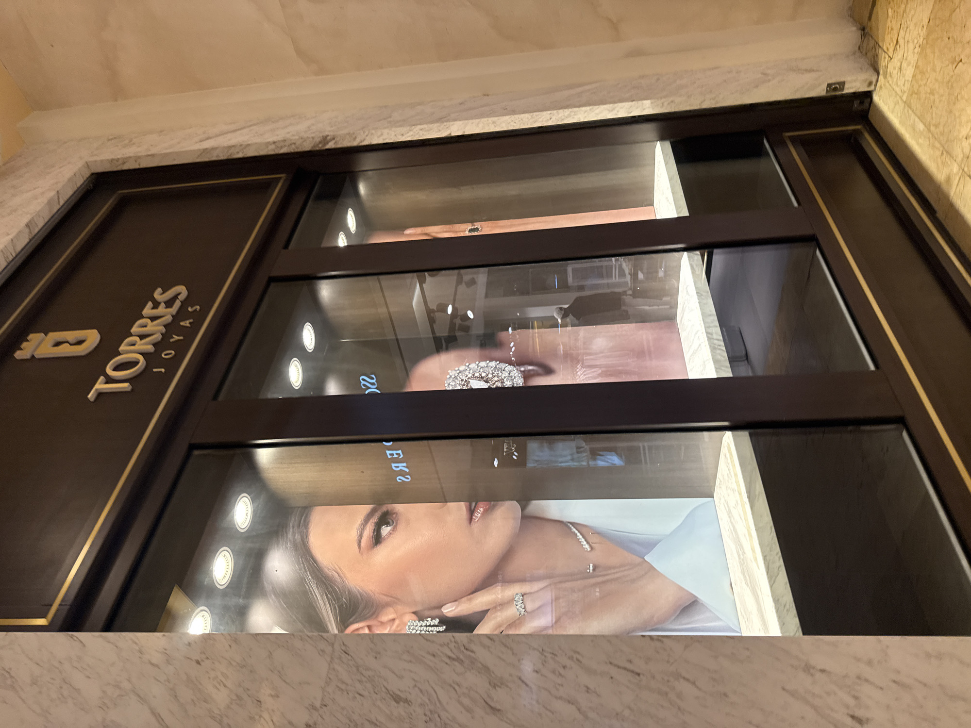 The image shows a store window of a jewelry shop named "TORRES JOYAS". The store's sign, which has the brand name in gold lettering over a dark brown background, is on the left. The window is displaying jewelry advertisements and actual jewelry pieces. A woman is featured in the advertisements. The store seems to be located in Puebla, Mexico. The shop facade is marble.
