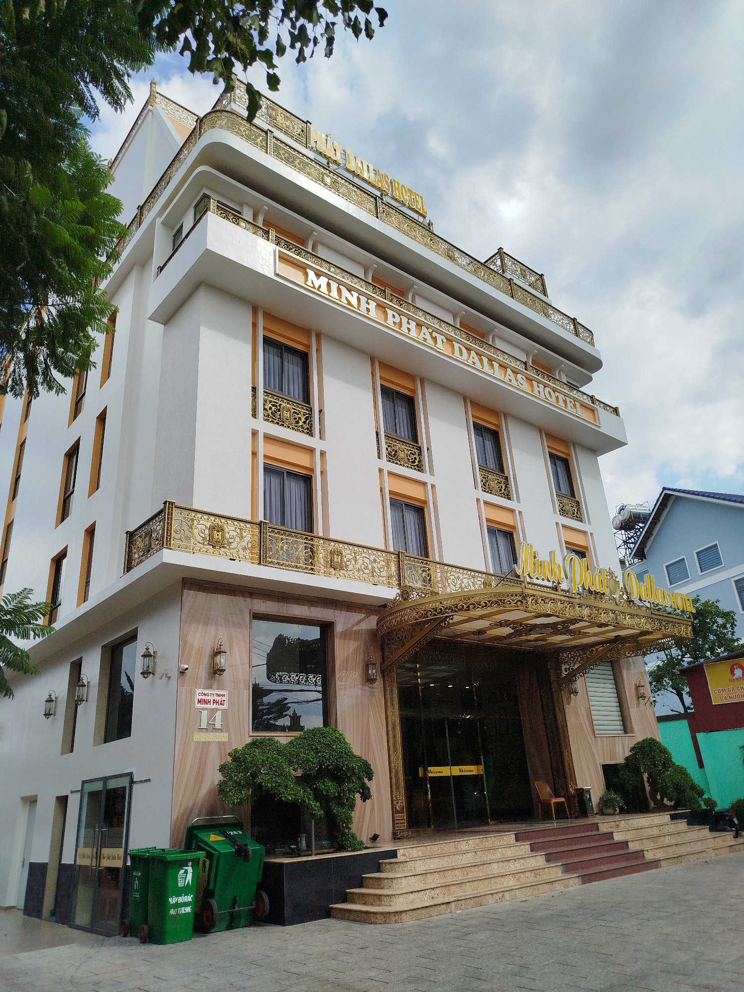 The image depicts the Minh Phat Dallas Hotel in Lâm Viên - Đà Lạt, Vietnam. The hotel is a multi-story white building with gold accents and balconies with ornate gold railings. The front entrance has a large gold canopy and multiple steps leading up to it. There is a sign on the front of the building and smaller signs on the entrance doors. The street in front of the hotel appears to be paved, and there are some trees and greenery around the building, including two green wheelie bins. To the right of the hotel's entrance, one may notice a neighboring building. The sky is overcast with clouds.