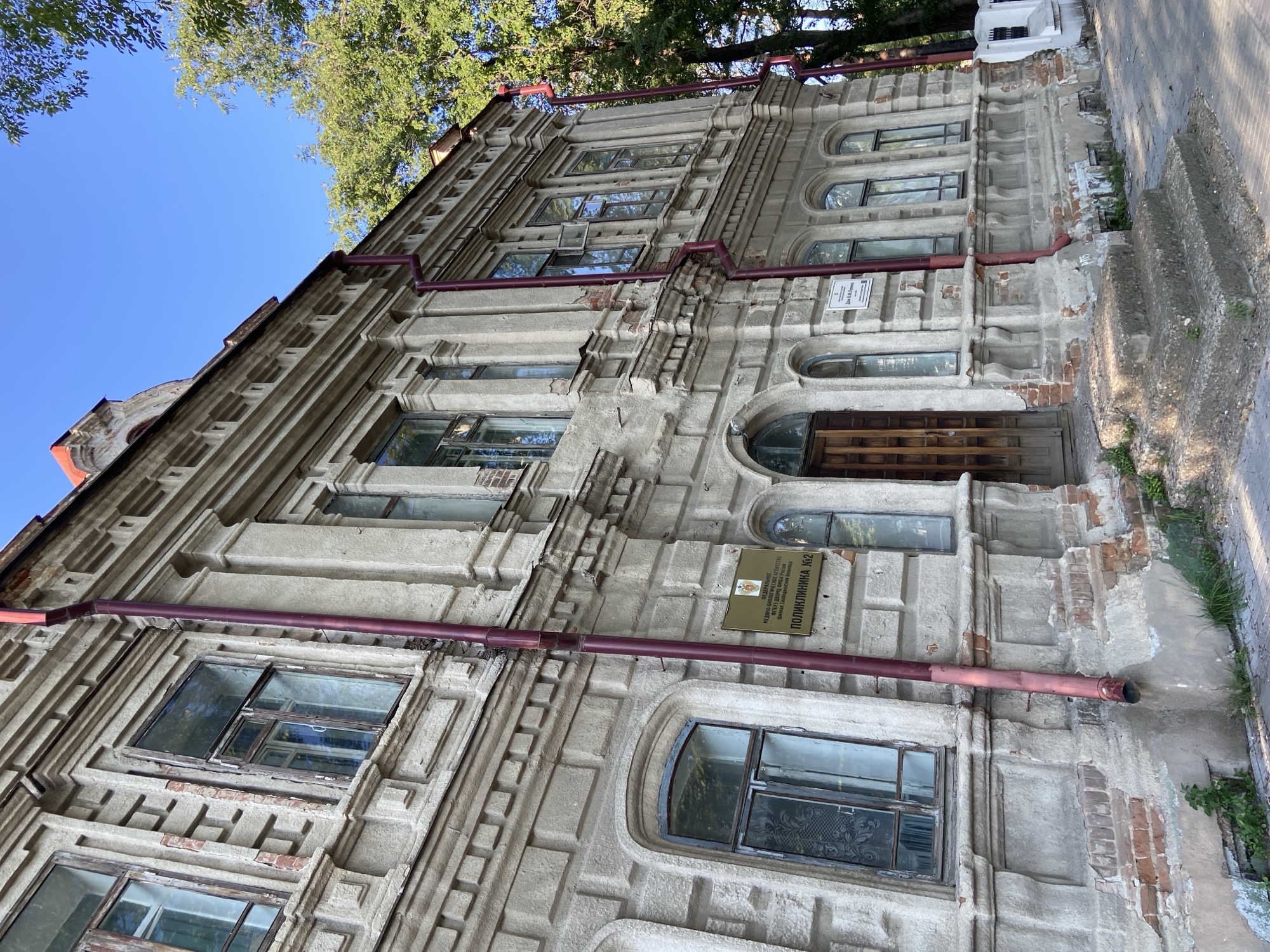 The image shows the facade of an old building in Blagoveshchensk, Russia. The building is constructed of stone and has several windows and a large wooden door. A sign on the building reads "ПОЛИКЛИНИКА №2" (Polyclinic No. 2). The building appears to be in a state of disrepair, with some of the plaster crumbling and bricks exposed. Trees are visible on the right side of the image, and the sky is clear and blue. The building is a historical site, formerly part of the estate of A. V. Lyukshem.