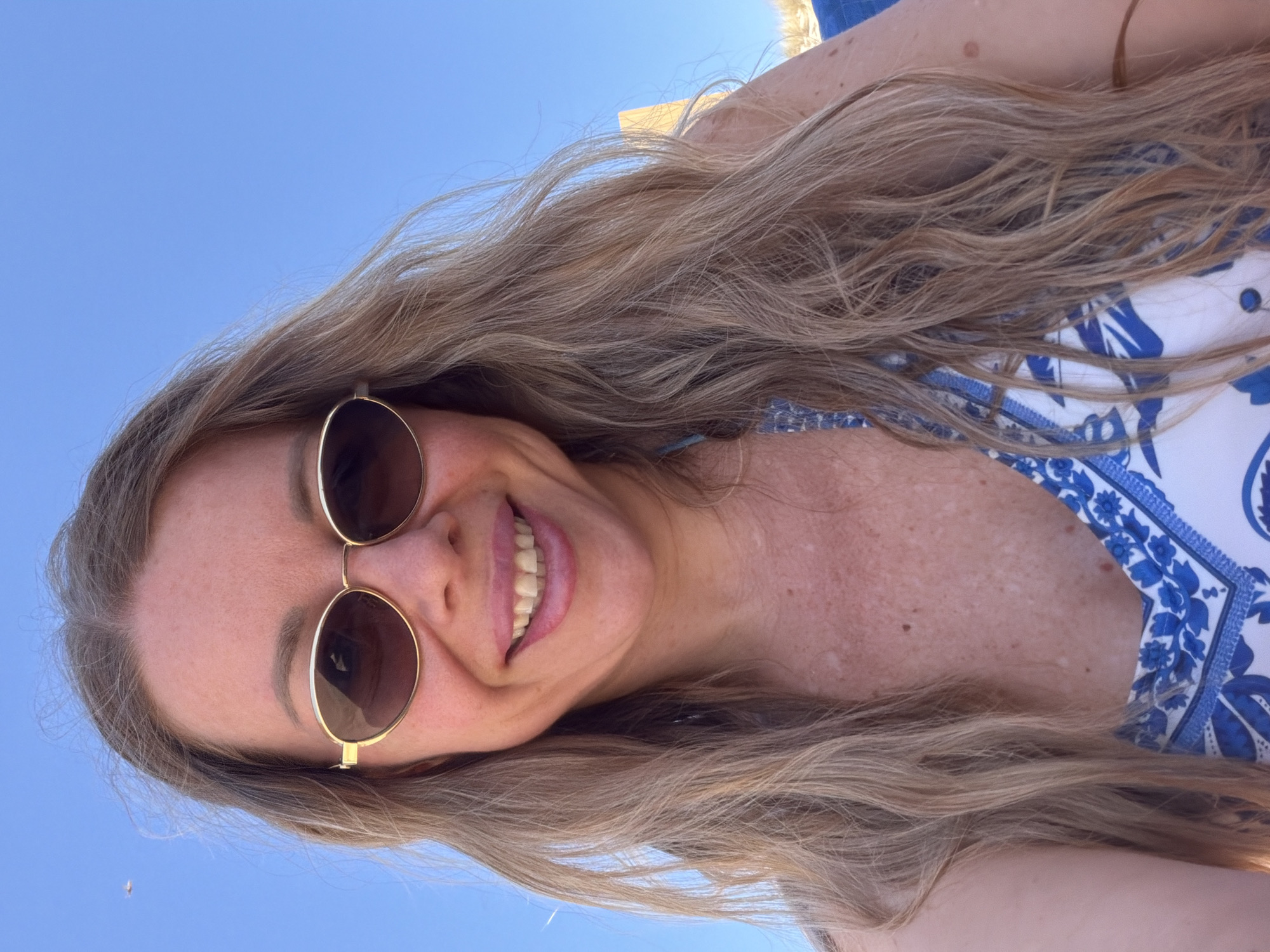 A medium shot portrait of a woman with long, blonde, wavy hair, wearing gold-rimmed oval sunglasses and a white and blue patterned bikini top. She is smiling with her mouth open, showing her teeth. The background is a clear, bright blue sky with a small, distant airplane visible in the upper left corner. The woman is positioned in the lower half of the frame, with her hair framing her face and falling over her shoulders. The image appears to have been taken outdoors, likely on a sunny day, with no discernable specific location within a city or country.