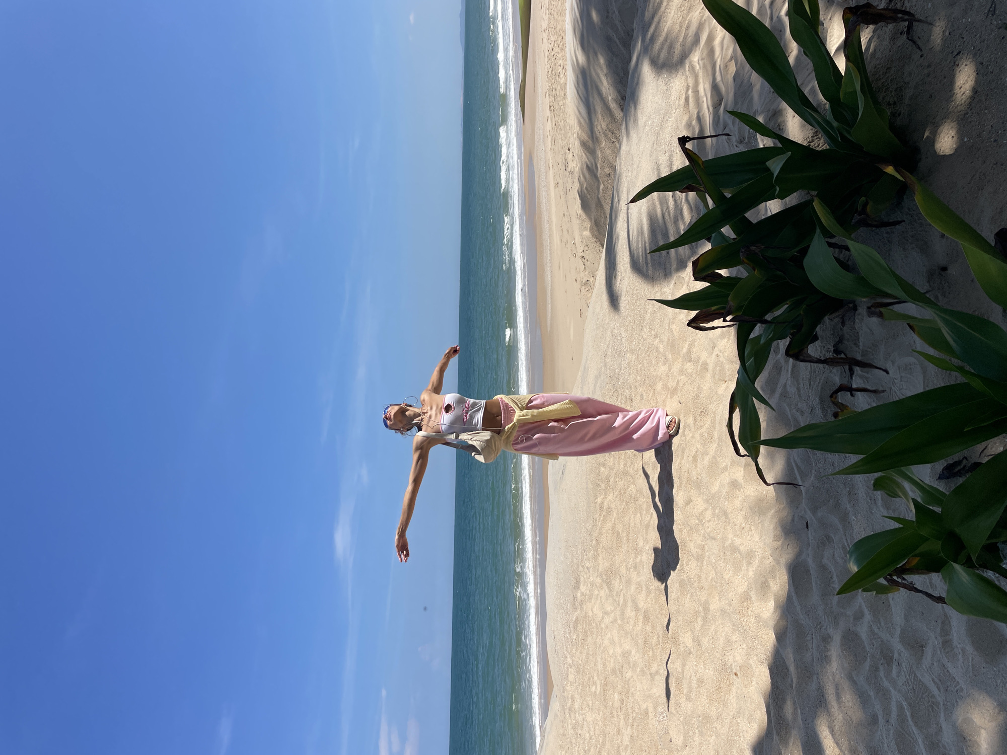A young woman with blonde hair, wearing a white cropped top, pink pants, and sunglasses, stands on a sandy beach in Phu Thuy Ward, Vietnam, with her arms outstretched towards the ocean. She is wearing a light-colored crossbody bag and a yellow garment tied around her waist. The sea is visible in the background, with waves gently lapping the shore under a clear blue sky. In the foreground, lush green plants cast shadows on the sand. The overall scene conveys a sense of freedom and relaxation in a tropical environment.