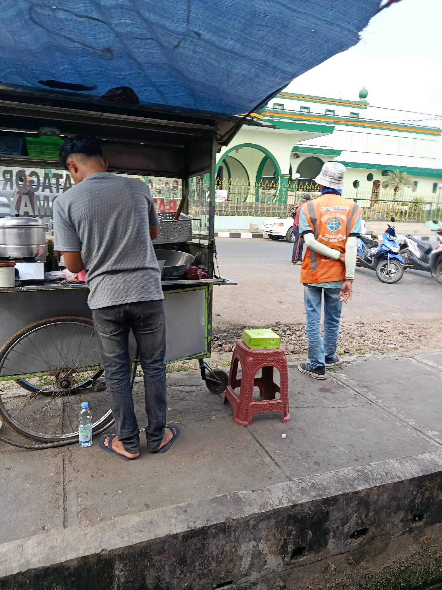 In Bululawang, Indonesia, a street vendor prepares food at a cart under a blue tarp. The cart is equipped with a steamer and various containers. A man in a grey striped shirt and dark jeans stands behind the cart, seemingly engaged in cooking. To the right, a person wearing a bright orange vest with reflective stripes and a white bucket hat stands with their arms behind their back, holding a cigarette. The person in the vest is facing away from the cart. In the background, a white and green mosque with arched windows and a dome is visible. Motorcycles and a white car are parked on the street. The scene is set on a concrete sidewalk adjacent to a road.