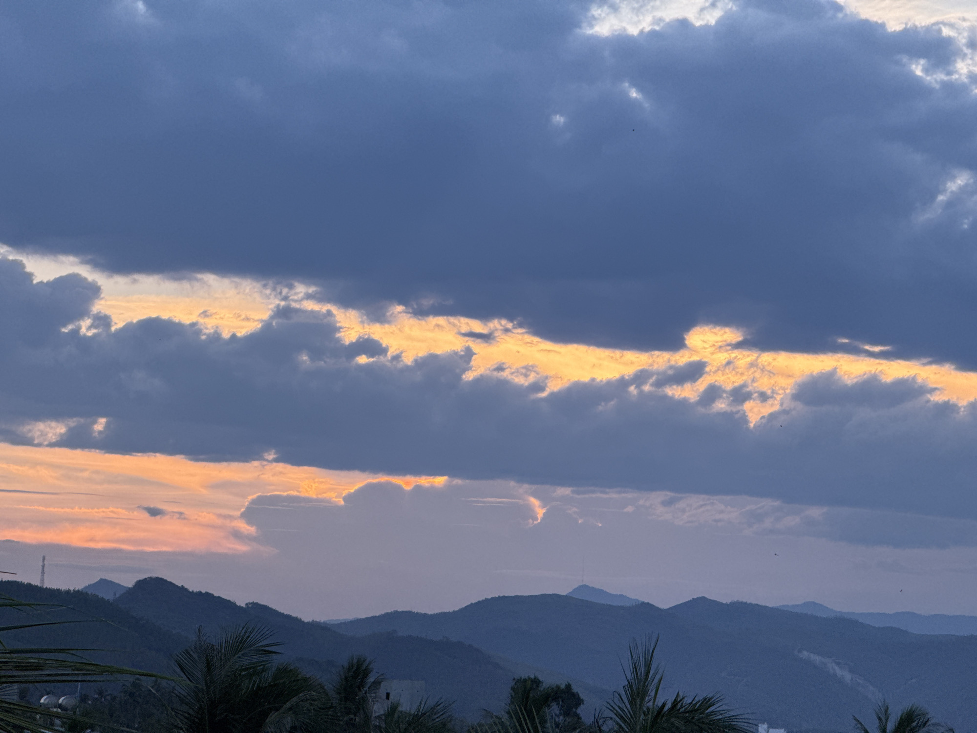 The image depicts a twilight scene over a mountainous landscape. The sky is filled with dramatic clouds, with a band of vibrant orange and yellow light breaking through the darker, purple-grey clouds near the horizon. These illuminated clouds suggest the sun is setting or has just set. Below the sky, layers of dark, silhouetted mountains recede into the distance. In the foreground, the fronds of palm trees are visible, framing the lower portion of the image. The overall atmosphere is serene and natural, with no people or specific structures clearly identifiable beyond distant hints of infrastructure like a radio tower.