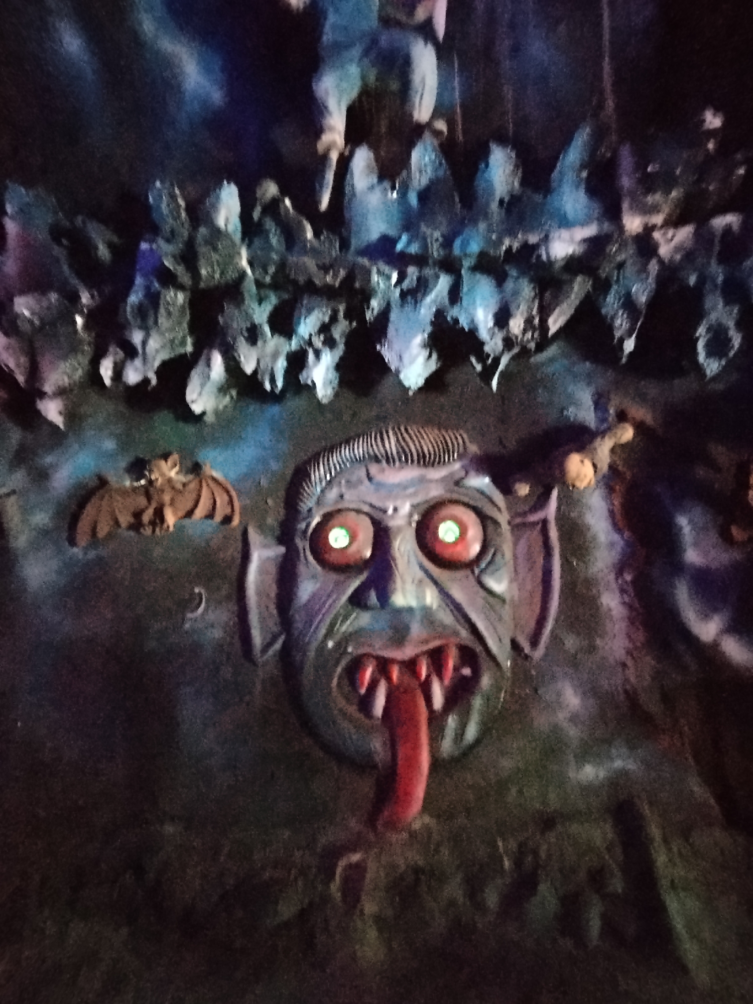 The image shows a wall decoration, seemingly inside a dark ride or haunted house attraction. The decoration consists of a menacing mask with glowing red eyes, a protruding tongue, and sharp teeth. To the left of the mask is a bat figurine, and above it is a rock-like structure, possibly meant to resemble stalactites. The wall surrounding the mask is dark and textured. The location is Păng Tiêng 1, Vietnam.