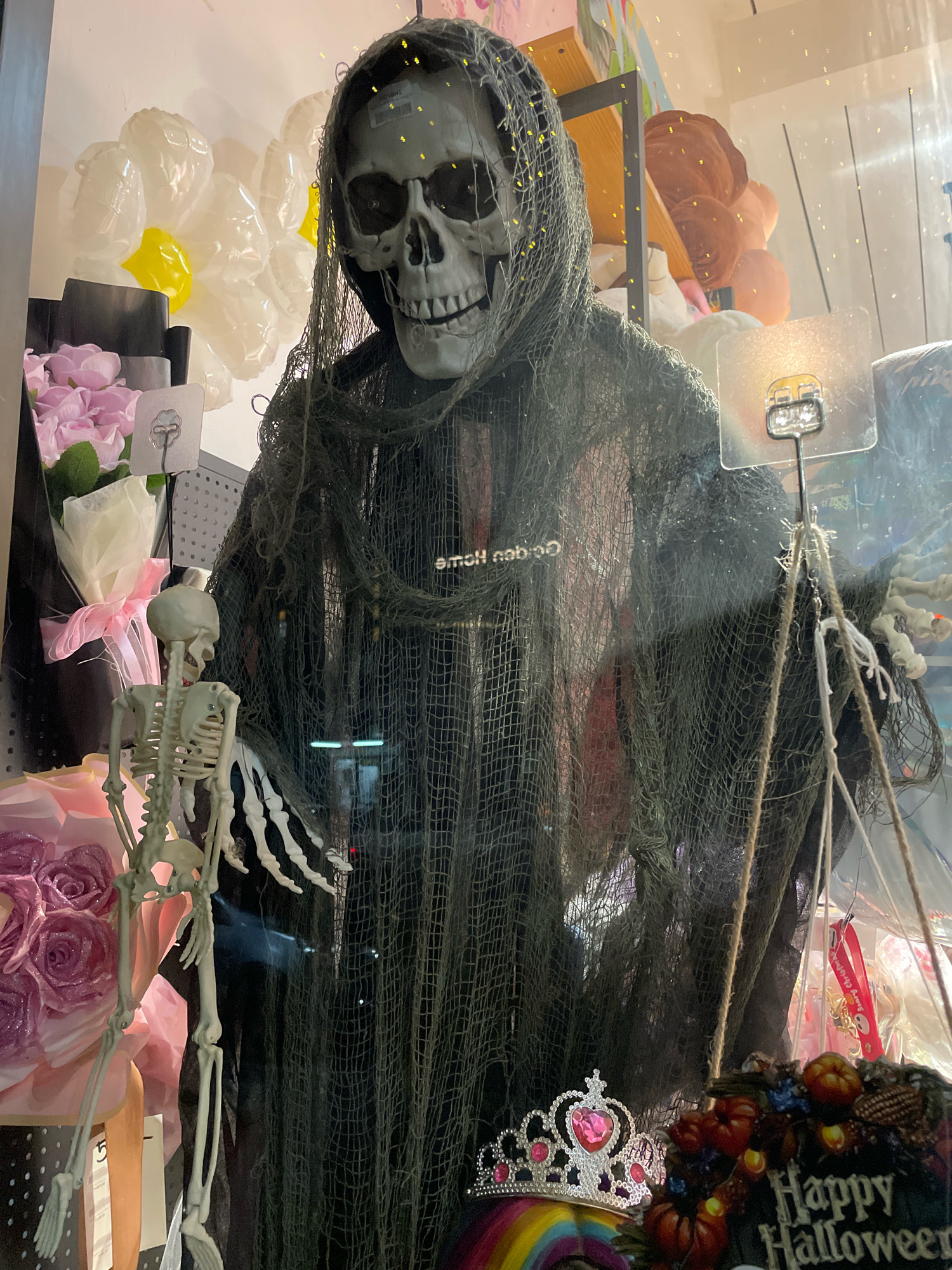 A Halloween-themed display is set up in a shop window in Pattaya City, Thailand. The central figure is a large grim reaper decoration with a skeletal head and a tattered black robe. To the left of the grim reaper, a smaller skeleton decoration is positioned. The display includes various Halloween-themed items such as balloons, artificial flowers, a tiara, and a "Happy Halloween" sign with miniature pumpkins. The overall scene suggests a commercial establishment preparing for the Halloween season.