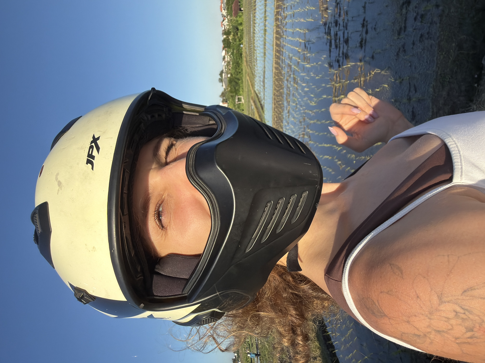 A woman wearing a helmet with the logo "JPX" is captured at eye-level, likely taking a selfie. She is positioned against the backdrop of a rice paddy field, reflecting the sky, indicating a rural area in North Kuta, Indonesia. Her right hand is raised slightly, the palm facing the viewer. She is wearing a brown and white tank top.