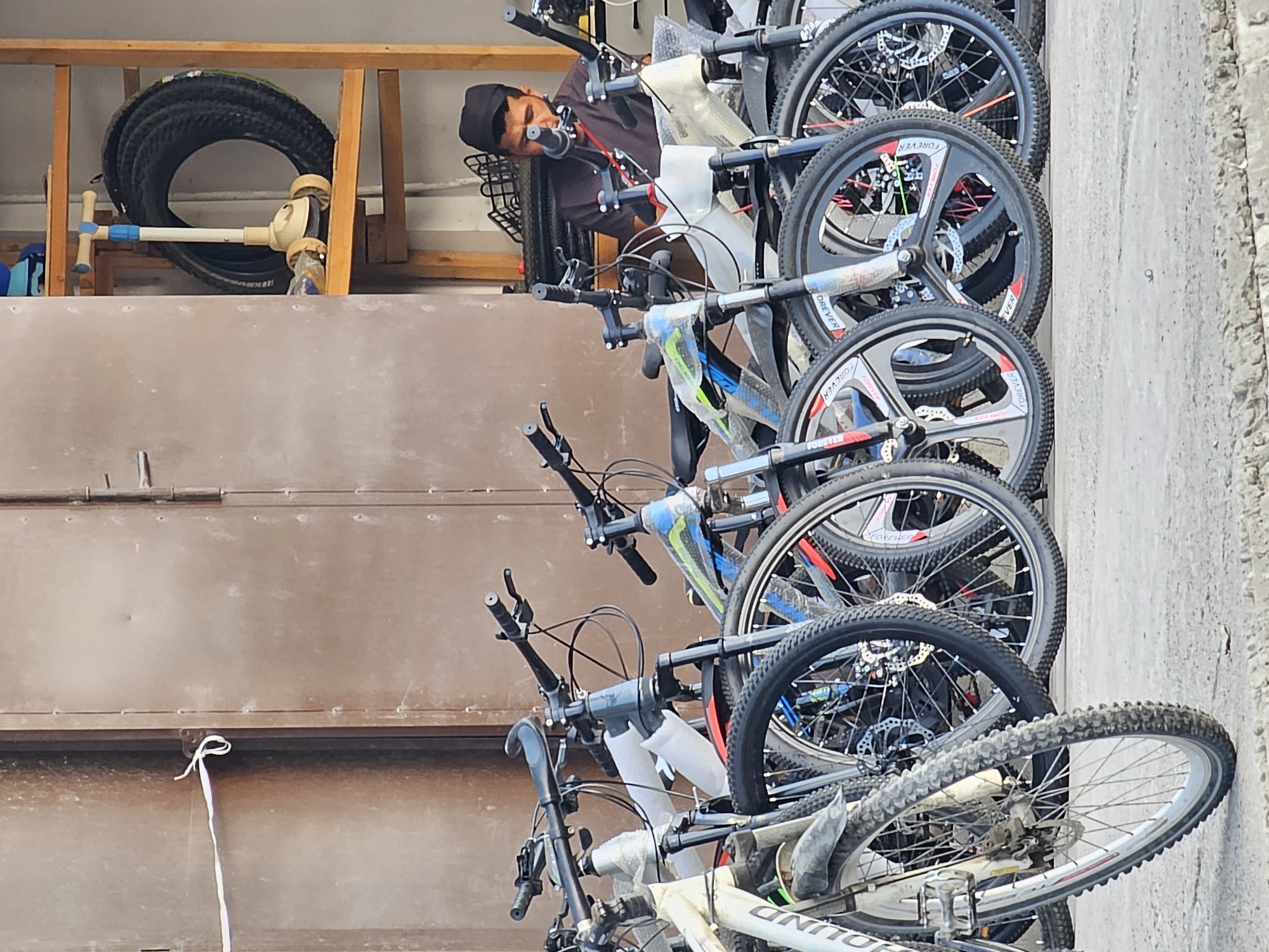 This image depicts a street-level view of a bicycle shop in the city of Özgön, Kyrgyzstan. In the foreground, several bicycles are lined up, their wheels and handlebars partially visible. The bikes appear to be new or for sale, with some still wrapped in protective plastic. The backdrop is a large, weathered metal door. To the right, a man wearing a black cap and dark shirt is visible behind the bicycles, seemingly attending to his inventory or customers. He is looking downwards, focused on something in front of him. On a wooden shelf behind the man, a coil of black hose and a child's scooter are stored. The ground appears to be paved with concrete. The scene suggests an outdoor commercial area, likely a sidewalk or entrance to a shop.