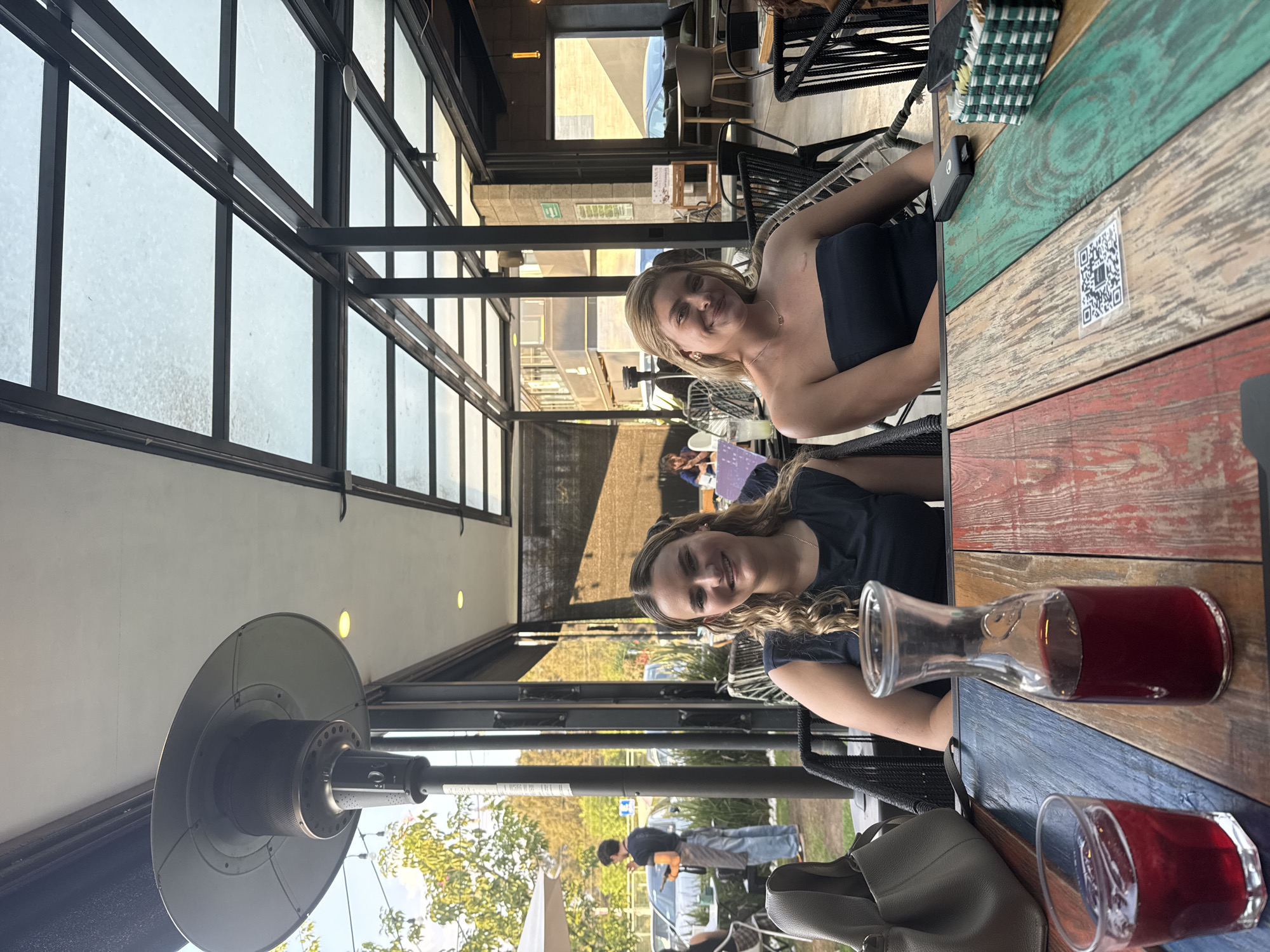 Two women are sitting at an outdoor table in San Bernardino Tlaxcalancingo, Mexico. The table is made of colorful wooden planks. A glass carafe filled with a red liquid and a matching glass with the same liquid are on the table. One woman is fair-skinned with blonde hair and is wearing a strapless top. The other woman has long, wavy brown hair and is wearing a dark-colored top. Behind them is a metal-framed glass structure that serves as the restaurant's outdoor seating enclosure. Outside the enclosure, trees and people are visible. A patio heater is positioned on the left. The sky is bright, suggesting it is daytime.