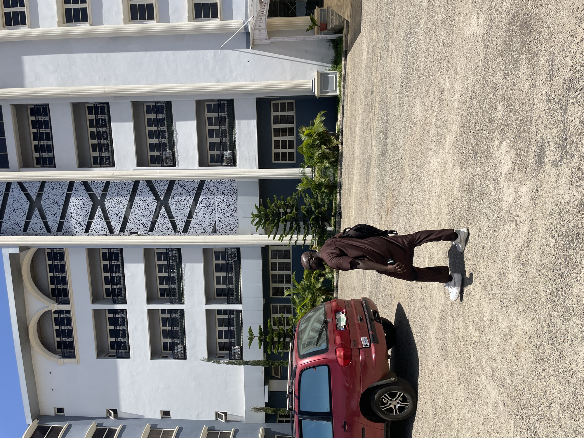 A man in a brown robe and white sneakers stands in a paved parking lot next to a red hatchback car. The lot is in front of a large, multi-story building with numerous windows and decorative architectural features. The building is located in Jos, Nigeria. The man is wearing a backpack and holding an object in his left hand. Vegetation, including trees and bushes, is visible between the building and the parking lot. The sky is clear.