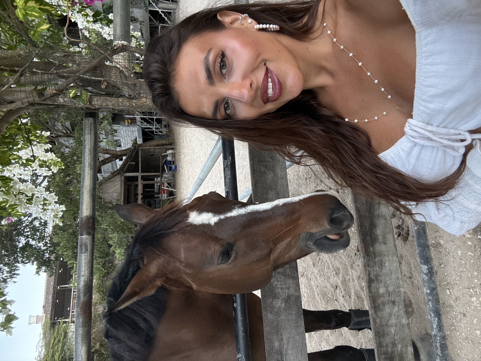 A woman with long dark hair, wearing a white off-the-shoulder top and multiple pearl necklaces and earrings, smiles while taking a selfie. She stands with a bay horse, distinguished by a white blaze on its face and dark leg wraps on its forelegs, positioned behind a wooden and metal fence. The horse appears calm, with its eye partially closed. The ground is sandy or dirt-like, and the background features lush tropical vegetation, including flowering bougainvillea with white and pink blossoms. In the distance, several buildings are visible, including a multi-story structure with a tiled roof and an open-air balcony, and another with grey walls and external stairs, characteristic of an equestrian facility or resort. This scene is located in Kuta Selatan, Indonesia.