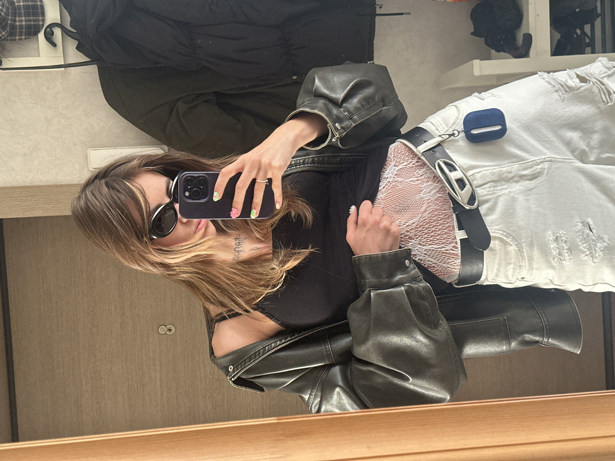 A young woman, with long blonde hair and sunglasses, is taking a mirror selfie. She is wearing a black tank top, a dark leather jacket, white ripped jeans, and a black belt with a silver buckle. White fishnet material is visible under her tank top, around her midriff. A small blue case is attached to her belt. The background appears to be an interior, possibly a room in an apartment, with a wooden door on the left and a mirror on the right reflecting the scene. A dark jacket is hanging on a rack in the background. The overall setting suggests a casual, indoor environment. There is no indication of the city of Moscow, Russia.