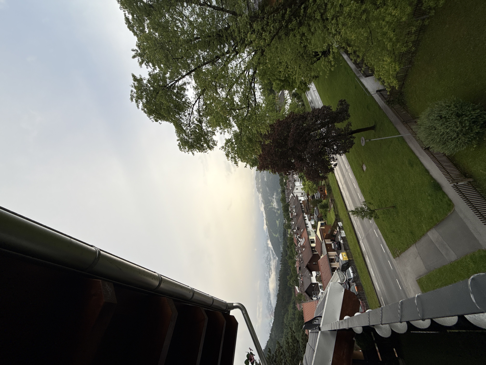 The image depicts a view from a balcony overlooking a street and houses in Garmisch-Partenkirchen, Germany. In the foreground, a dark wooden soffit and a metal gutter are visible. The scene extends to a paved road with parked cars and houses with tiled roofs. Beyond the houses, a landscape of green hills and distant mountains shrouded in mist is visible under an overcast sky. The overall impression is a residential area within a mountainous region.