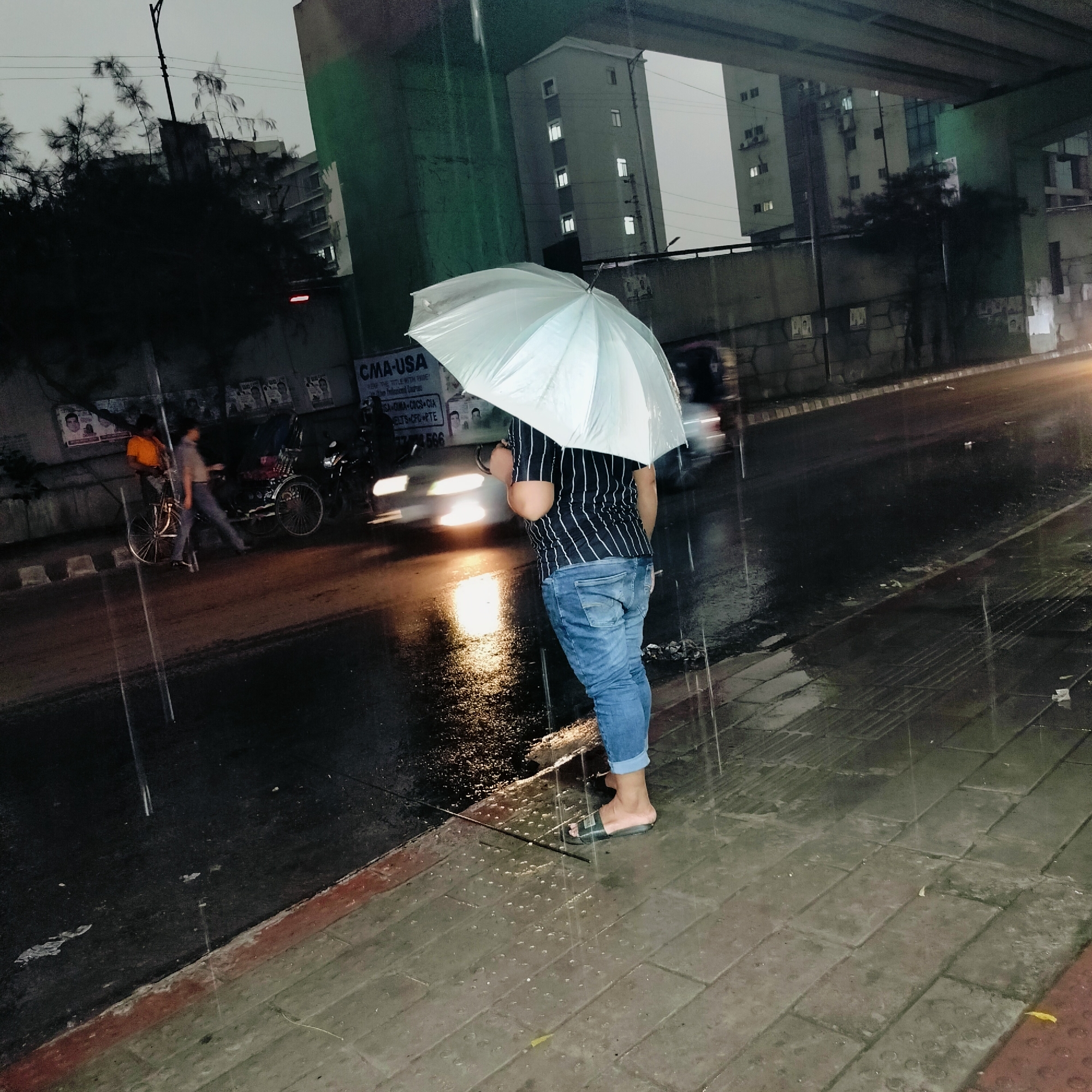 A man with an umbrella stands on a wet sidewalk in Dhaka, Bangladesh. The man is wearing jeans and a striped shirt, and he is holding a clear umbrella. Cars drive past on the wet road behind him, their headlights reflecting in the puddles. In the background, there are buildings and an elevated overpass. It is raining. A sign that reads "CMA-USA" is visible in the background, along with Bengali script and other posters. Two men with bicycles are visible on the left side of the image. The overall tone of the image is somber and wet.