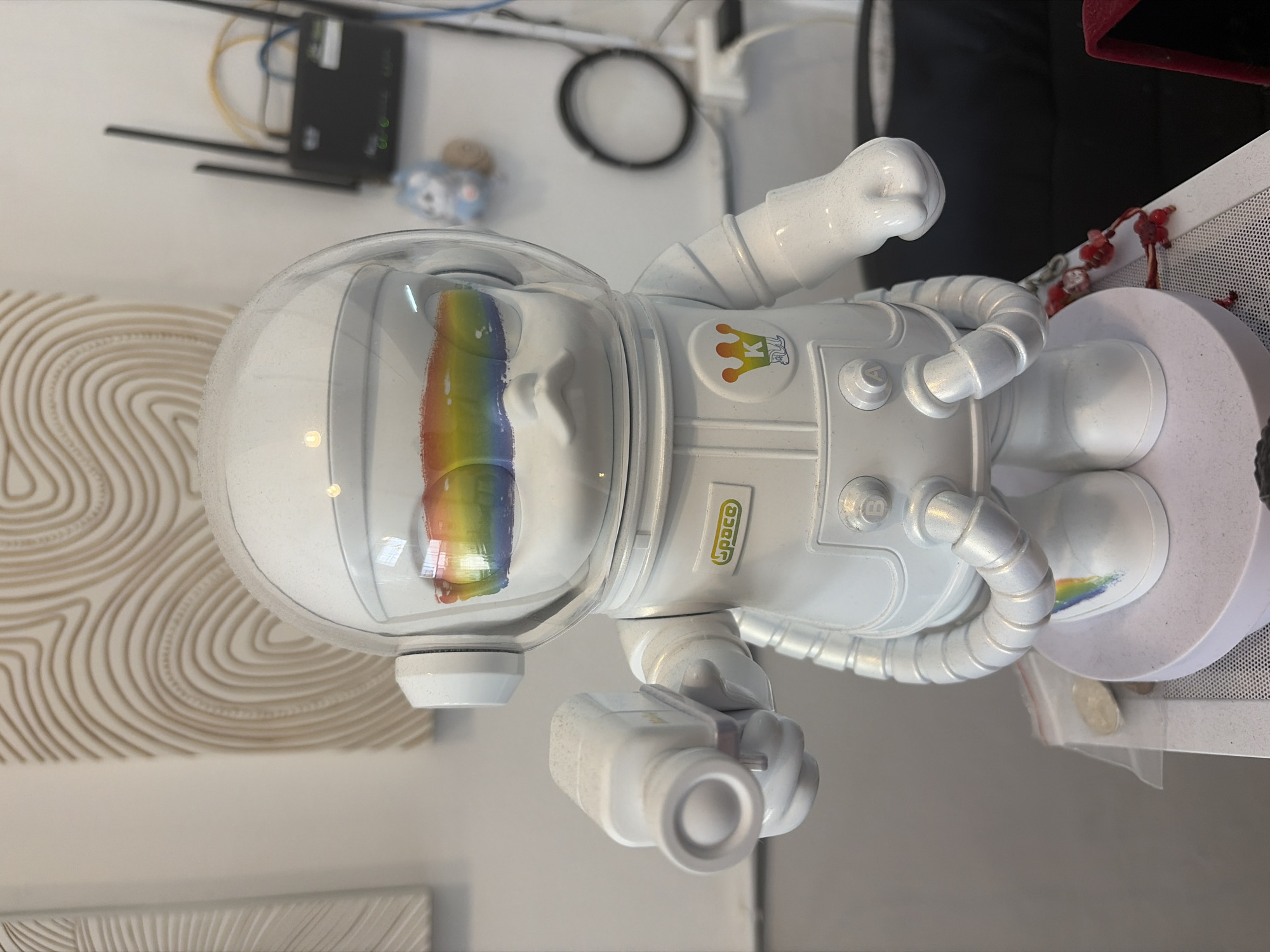 A white toy astronaut figure stands on a white circular base. The figure has a clear helmet with rainbow-colored markings visible inside, suggesting eyes or a visor decoration. It holds a white device that resembles a camera or a ray gun in its left hand. The astronaut figure wears a suit with various details, including a "space" logo on its chest and a crown emblem with a "K" inside. The background includes a textured wall art, a black and white router with antennas, and a small blue and white cat figurine. There are also some wires and a power outlet visible. The overall scene appears to be indoors, possibly in a shop or a home in Bangkok, Thailand.
