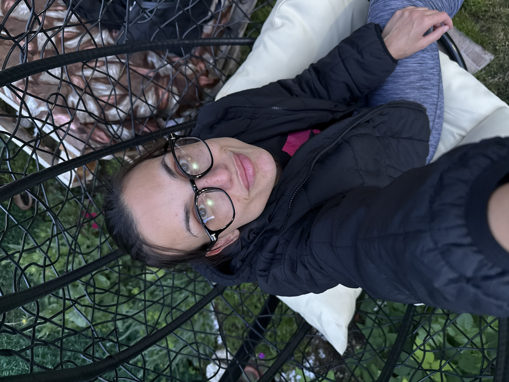 A woman wearing glasses and a black puffer jacket is lying in a black metal hanging chair with white cushions. She is wearing blue leggings. The chair is surrounded by green foliage. The location is unknown, and there is no information indicating it is in Pleven, Bulgaria.