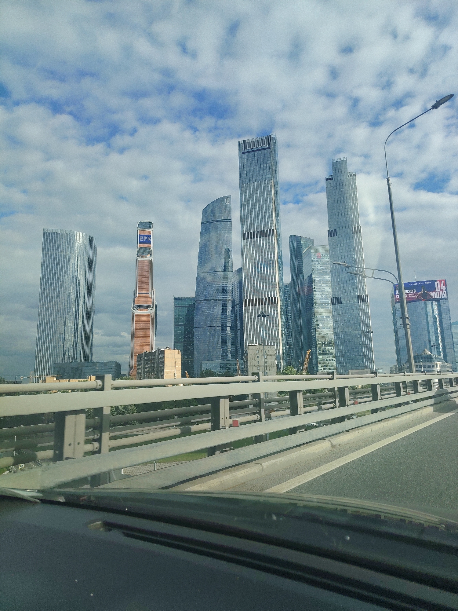 The image shows a view from a vehicle traveling on a highway, looking towards a modern cityscape in Moscow, Russia. Dominating the skyline are several very tall, contemporary skyscrapers with glass facades. The tallest and most prominent is the tower with the blue "BPK" logo at the top. To its left is a very tall, slender cylindrical building. To the right of the "BPK" tower are several other skyscrapers of varying heights and designs, some with distinctive angled tops. In the foreground, a highway barrier and the road are visible. The sky is partly cloudy with patches of blue. The overall impression is one of urban development and modern architecture in Moscow.