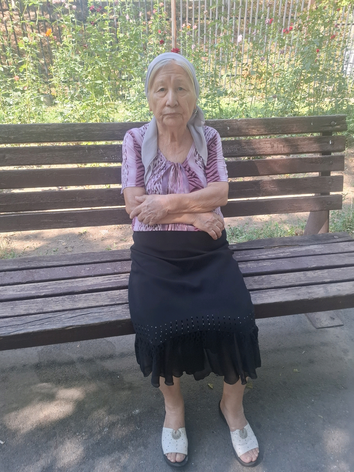 An elderly woman is seated on a wooden park bench, positioned centrally in the frame. She is wearing a pink and purple patterned blouse, a light gray headscarf, and a black skirt with a decorative hem. Her arms are crossed, and she is looking directly at the camera with a neutral expression. The bench is weathered and appears to be in a park setting, with a dense green bush with some red and orange flowers behind her, and a metal fence behind the bush. The ground in front of the bench is paved with asphalt, showing patches of sunlight and shadow. The scene is likely outdoors, possibly in Almaty, Kazakhstan, given the context.