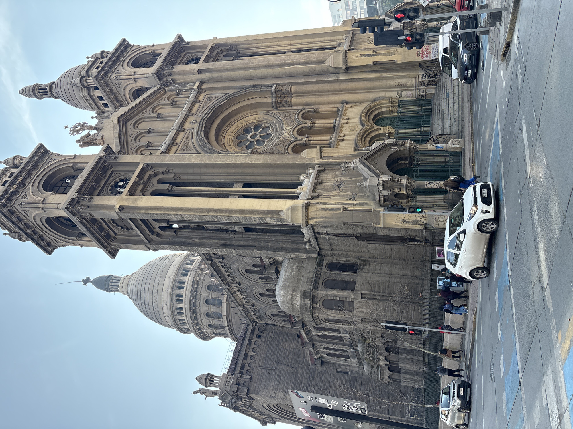 This is a medium shot, eye-level, exterior photograph of the Metropolitan Cathedral of Santiago, Chile, from a street intersection. The majestic, large stone cathedral dominates the right two-thirds of the frame, featuring an ornate facade with arched windows, columns, and two tall bell towers topped with domes. A large central dome with a smaller spire is visible behind the right bell tower. The cathedral's stone exterior appears aged, with some areas showing signs of weathering and graffiti.
In the foreground, a street intersection with pedestrian crosswalks is visible. Cars are present: a white Kia Picanto is driving away from the viewer towards the left, a dark grey Audi A7 is stopped at the intersection, and a red sedan is partially visible behind it. A white van is parked to the right of the Audi. Pedestrians are crossing the street or walking on the sidewalk. Several traffic lights are visible, with one showing a red signal and another showing a green signal. The sky is overcast and a pale blue. The overall scene captures a busy urban environment in Santiago, Chile, with the imposing presence of the cathedral as the central element.