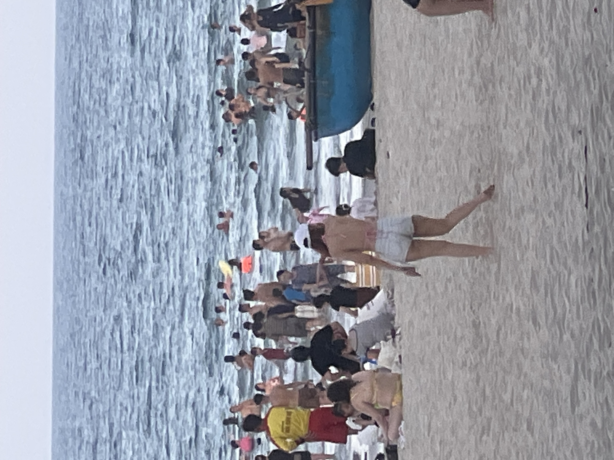 This media file depicts a crowded beach scene in Đà Nẵng, Vietnam. Numerous people are engaged in various beach activities, including swimming in the ocean, relaxing on the sand, and walking along the shoreline. The water appears choppy, suggesting it might be windy. The beach is sandy, with a line of people near the water's edge. In the background, a blue object, possibly a boat or a structure, is visible in the water. The sky is overcast, indicating a cloudy day, and the overall lighting suggests it could be late afternoon.
The image captures a lively atmosphere with many individuals enjoying their time at the beach. Some people are partially submerged in the water, while others are standing or sitting on the sand. A person in a yellow and red shirt, potentially a lifeguard, is visible among the crowd. The density of people suggests a popular tourist spot or a local gathering. The composition of the image is from a high vantage point, looking down on the beach and the sea. The focus is on the human activity and the expansive beach and ocean. No specific text is clearly legible in the image.