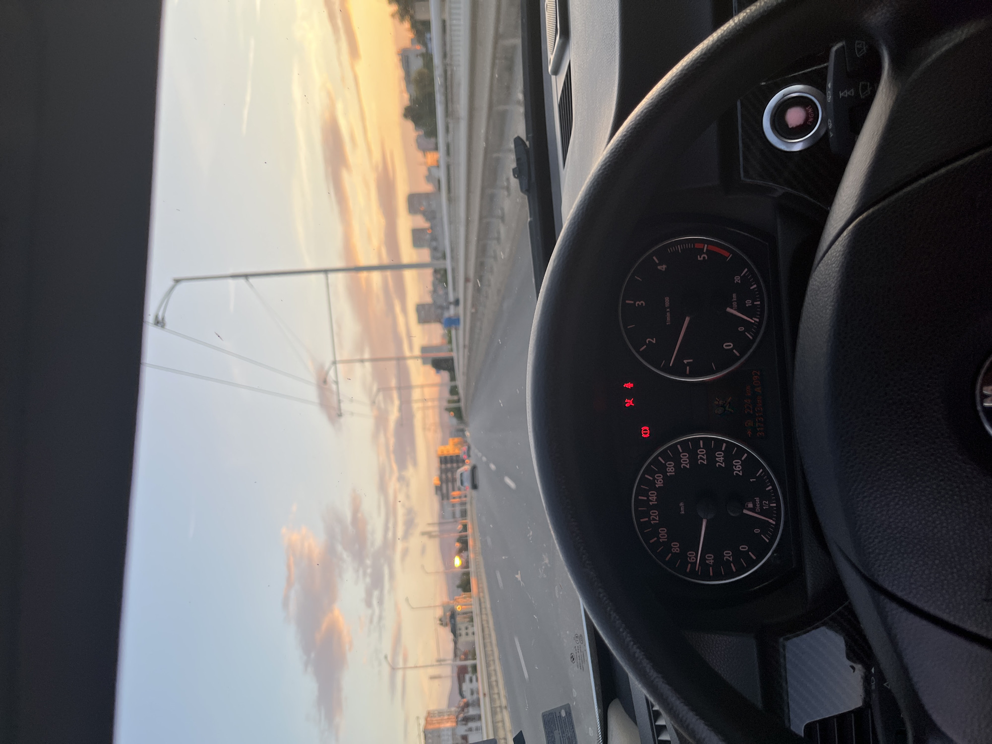 The image depicts a view from the driver's seat of a car, looking forward through the windshield. It is sunset, with the sky filled with soft orange and pink clouds. The car is on a multi-lane road, possibly a highway or a bridge, with streetlights and buildings visible in the distance, suggesting an urban environment. The dashboard of the car is visible in the foreground, showing the speedometer indicating approximately 100 km/h, and a tachometer. A red engine start/stop button is also visible. The overall impression is a drive through a city at dusk. The specific location in Sofia, Bulgaria, is not identifiable from the image alone.