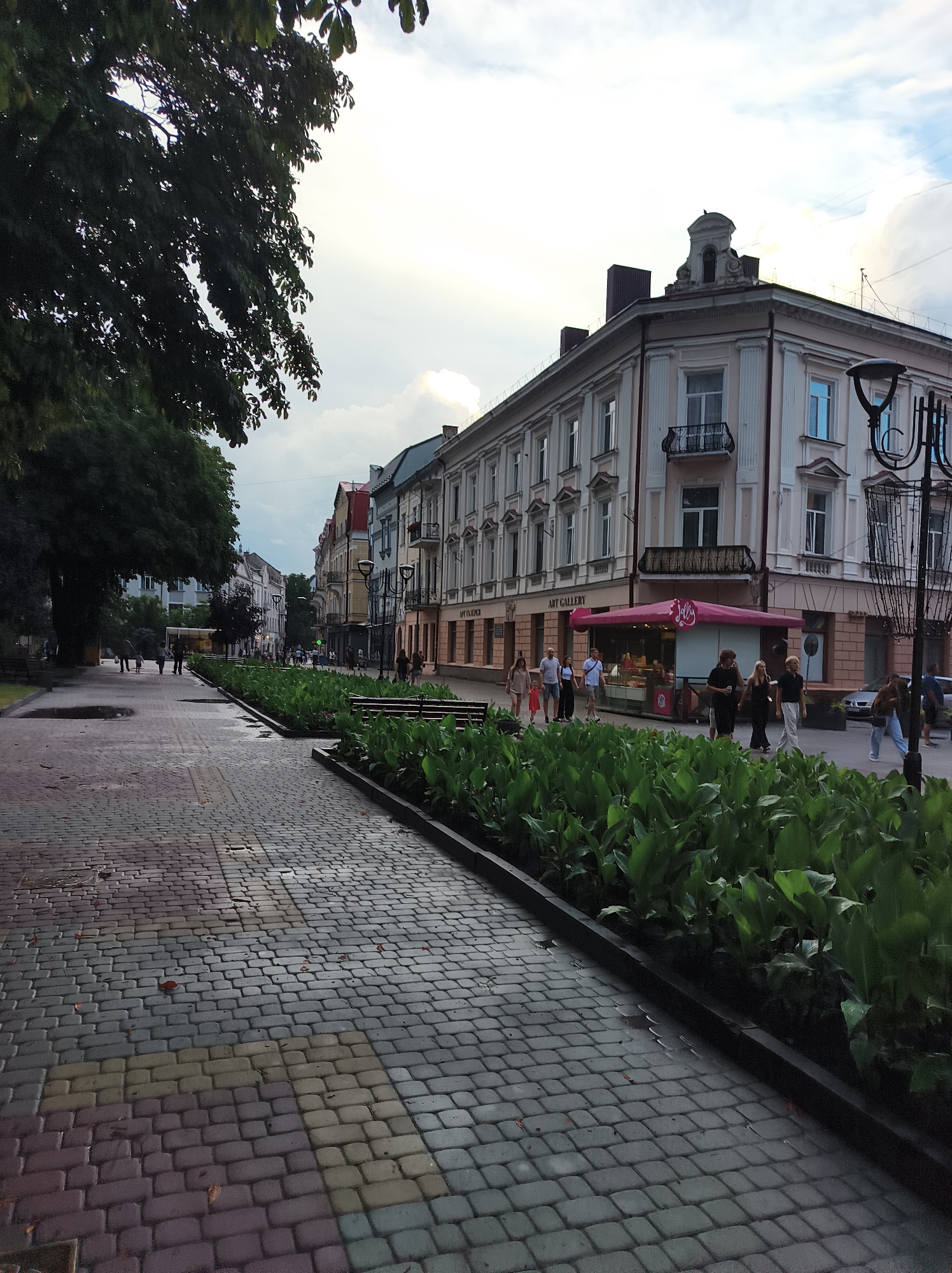 A pedestrian walkway paved with multicolored cobblestones is in the foreground, with a dark curb and lush green plants bordering it. Several people are walking on the walkway, some from the left and some from the right. Across the walkway and the planted area, a street lined with historic buildings stretches into the distance. These buildings feature architectural details like columns, balconies, and ornate cornices. One building has a prominent pink awning with the word "Jolly" written on it. The sky above is cloudy, with patches of lighter sky suggesting the time is either early morning or late afternoon. The scene appears to be in a European city, likely in Ternopil, Ukraine, given the architectural style and presence of pedestrian zones in urban areas.