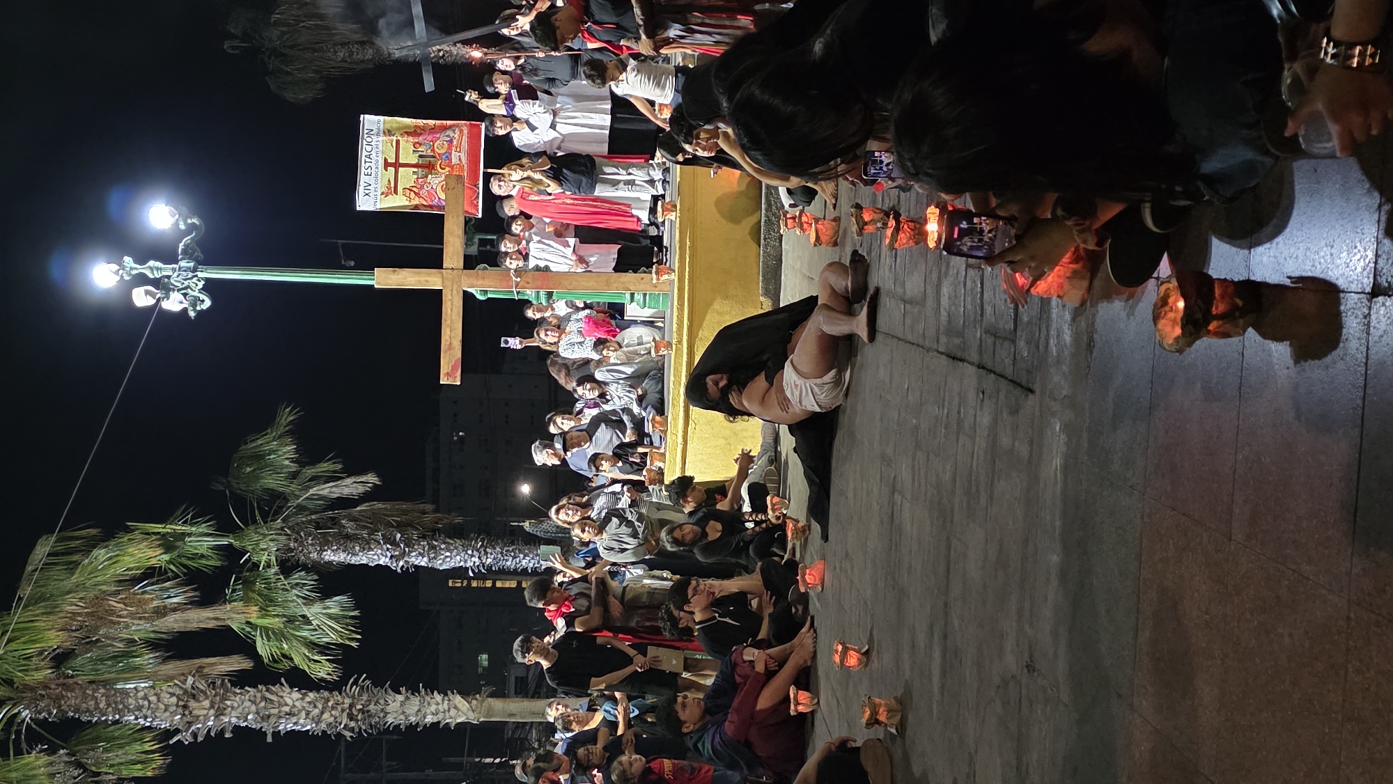 At night in an urban environment in Unknown, Unknown, a street performance depicting the crucifixion is underway. Actors in religious attire are gathered around a cross on a raised platform. A person lies on the ground being cradled, surrounded by an audience. Many people are holding candles and taking pictures with their phones. A lamp post and palm trees are also present.