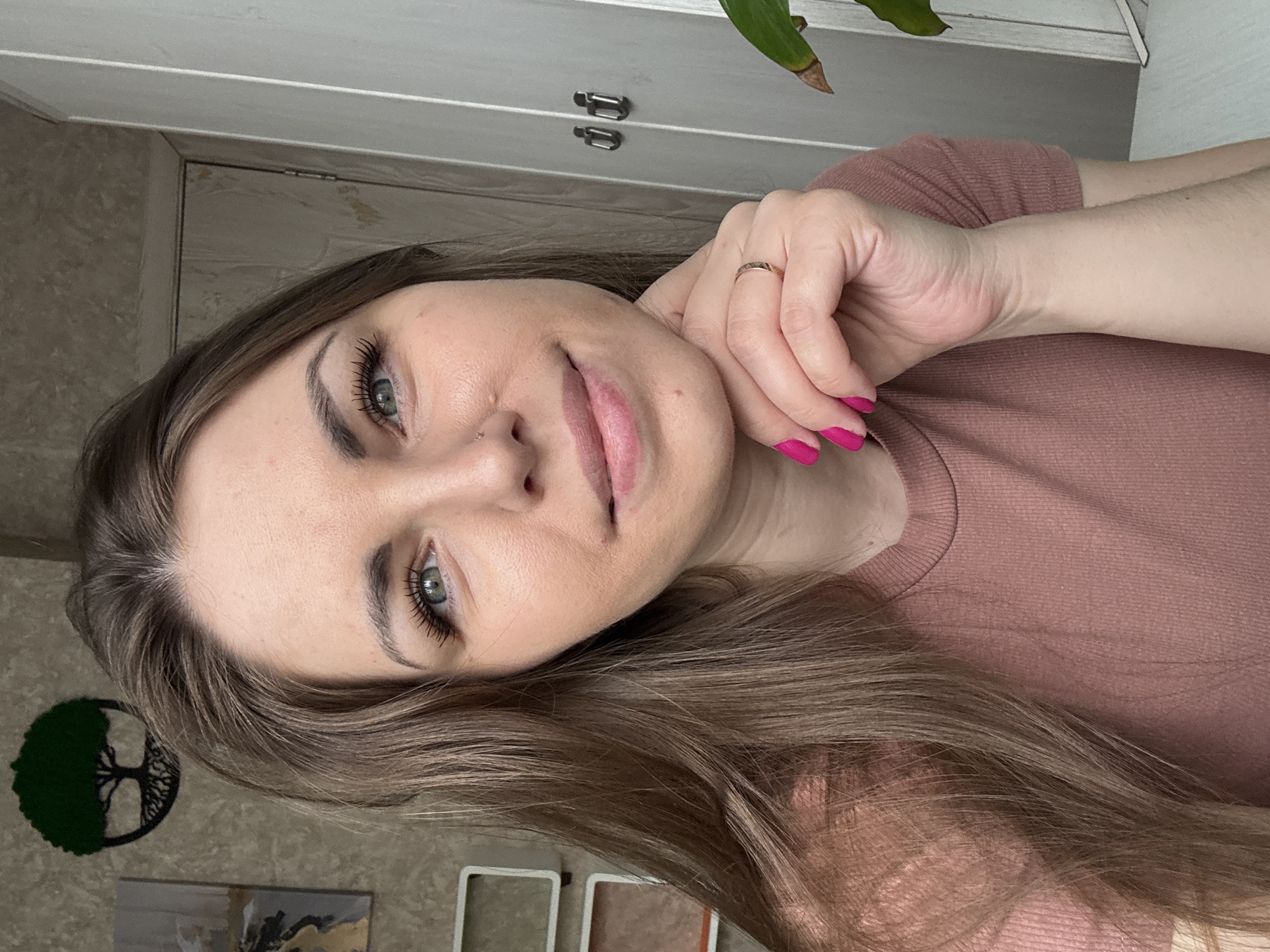 A selfie-style portrait features a woman with long, light brown hair, light-colored eyes (possibly blue or green), and defined eyebrows, gazing directly at the camera with a calm, subtle smile. She has a small stud piercing on her nose and her lips are slightly parted, revealing a light pink lip color. Her eyelashes appear long and curled. She wears a simple, ribbed pink or mauve top. Her right hand is positioned near her chin, with her fingers gently resting against her cheek. Her nails are painted a vibrant fuchsia pink, and a slender ring is visible on her ring finger.
The setting appears to be indoors, likely a home in Раздолье, Russia. In the background, above the woman's head, there is a light-colored cabinet or set of drawers with metallic handles. A portion of a green leafy plant is visible in the top right corner. To the left of the woman, a circular dark green "tree of life" wall decoration is visible. Below it, on the wall, there are small framed artworks, possibly abstract, with hints of gold and grey tones. Further down, small decorative boxes or frames rest on a surface. The overall lighting suggests daytime. There is no visible text in the image.