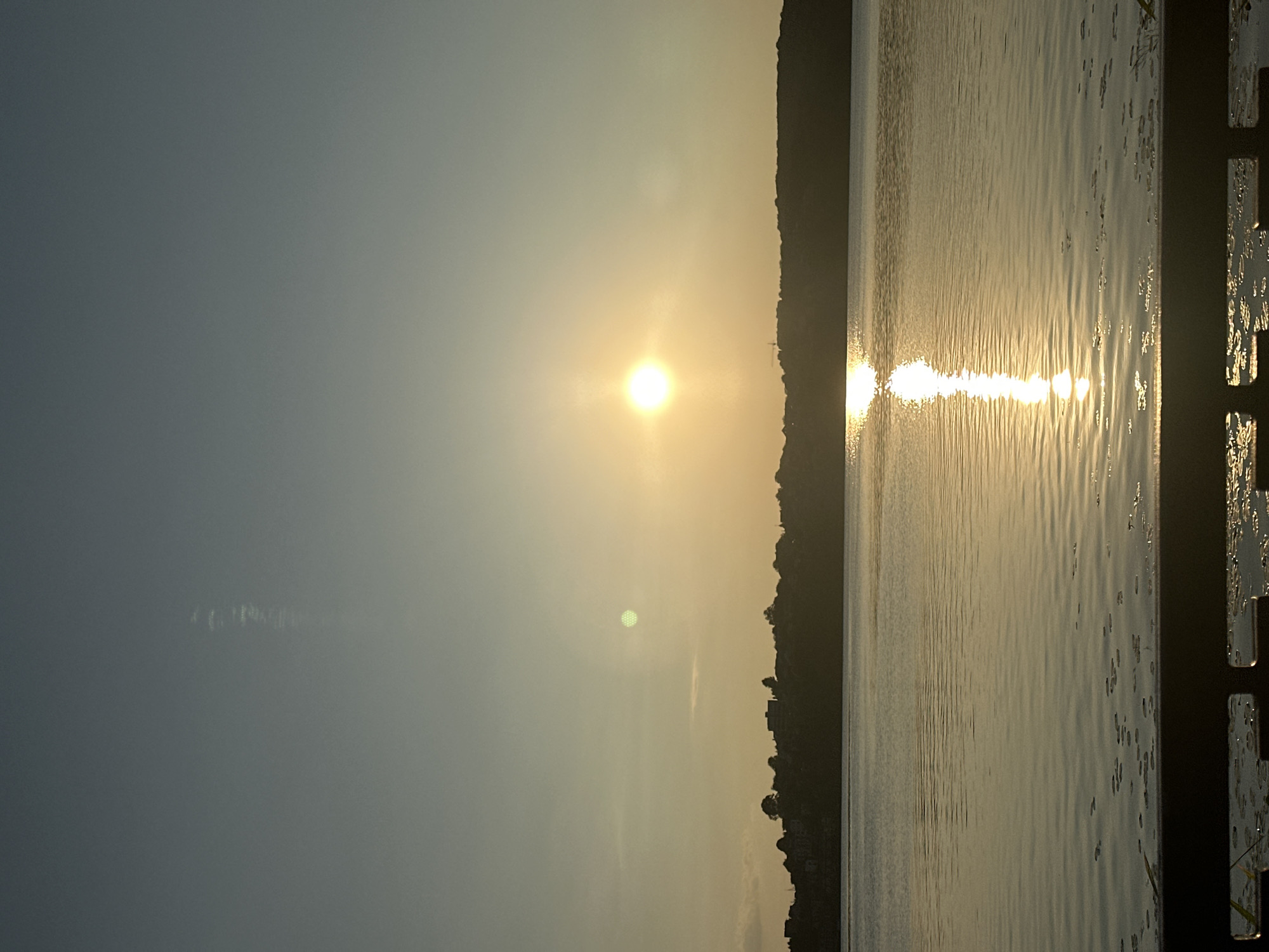 This image depicts a serene sunset over the Ternopil pond in Ternopil, Ukraine. The sun, a bright orb of light, is positioned in the upper right quadrant of the sky, casting a warm, golden glow across the water. Its reflection creates a shimmering path on the surface of the pond, drawing the eye towards the horizon. The distant shore is silhouetted against the bright sky, with the faint outline of buildings and trees visible. The water is calm, with gentle ripples disturbing its surface. In the foreground, a dark railing partially obstructs the view, suggesting a vantage point from a pier or promenade. The sky transitions from a clear, pale blue at the top to a soft yellow-orange near the horizon, indicating the late hour of the day. A faint, wispy cloud stretches vertically in the upper left corner of the sky. The overall atmosphere is peaceful and tranquil.