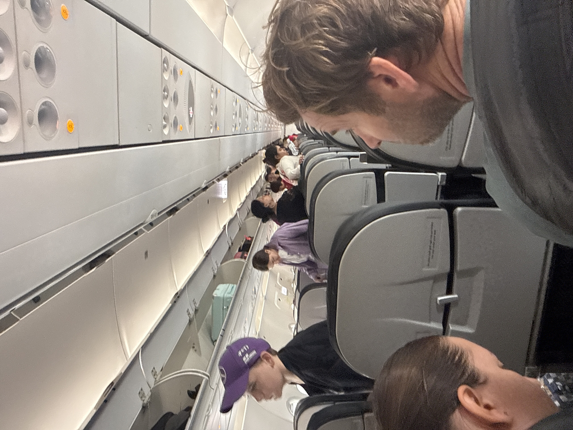 The image depicts the interior of an airplane cabin filled with passengers. The overhead compartments are visible along the side of the cabin. Several people are seated in the airplane seats, including a person wearing a purple baseball cap. The passengers are of various ages and wearing different colored clothing. The focus appears to be on the passengers and the overall scene inside the airplane cabin. There is no information related to Đà Nẵng, Vietnam, in the image.
