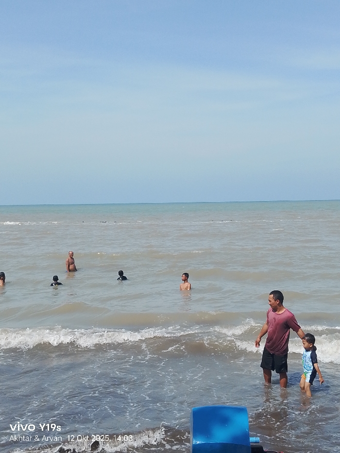 The image depicts a coastal scene on a clear, sunny day in a location identified as Limbangan, Indonesia. The upper half of the frame is dominated by a light blue sky with no visible clouds, meeting the ocean horizon. The ocean water is a murky, light brown or greyish-green color, with small waves breaking near the foreground shore.
Approximately seven to eight individuals are present in the water. In the immediate foreground, on the right side, an adult male with short dark hair, wearing a dark red t-shirt and dark shorts, stands in the water, which reaches his waist. He holds the left hand of a young child, who wears a multi-colored (blue, yellow, white) full-body swimsuit and is submerged to mid-thigh. Both face towards the right side of the frame.
Further out in the mid-ground, an adult male with light skin and thinning grey hair, shirtless, stands chest-deep in the water, looking to his left. To his right, an individual with a dark head and shoulders is partially submerged. Directly to the right of the shirtless man, another individual is submerged to the chest, facing slightly right. Two other individuals are visible further left, mostly submerged with only their heads and shoulders appearing above the water. All individuals are engaged in wading or swimming.
In the bottom right corner, a partial view of a bright blue, curved plastic object is visible, partially submerged in the water. The image includes a timestamp and device information in the bottom left corner: "vivo Y19s Akhta & Arvan 12 Okt 2025, 14:03".