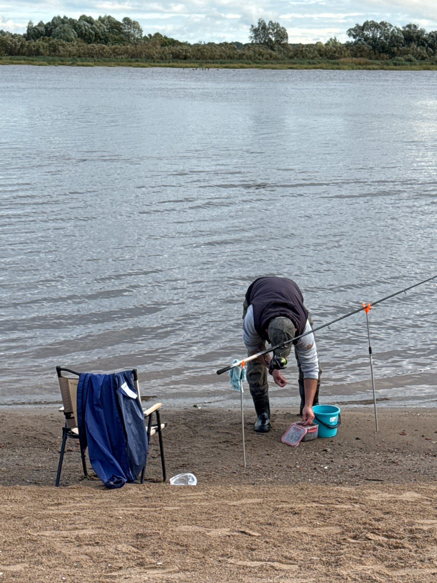 The image depicts a sandy riverbank with a person fishing. The person, wearing waders and a vest, is bent over attending to fishing gear. Nearby, a folding chair draped with a blue garment, a bucket, and a bait container are present. The river occupies the background, with a treeline and cloudy sky visible on the opposite bank. The scene is set in Veliky Novgorod, Russia, along the Volkhov River.