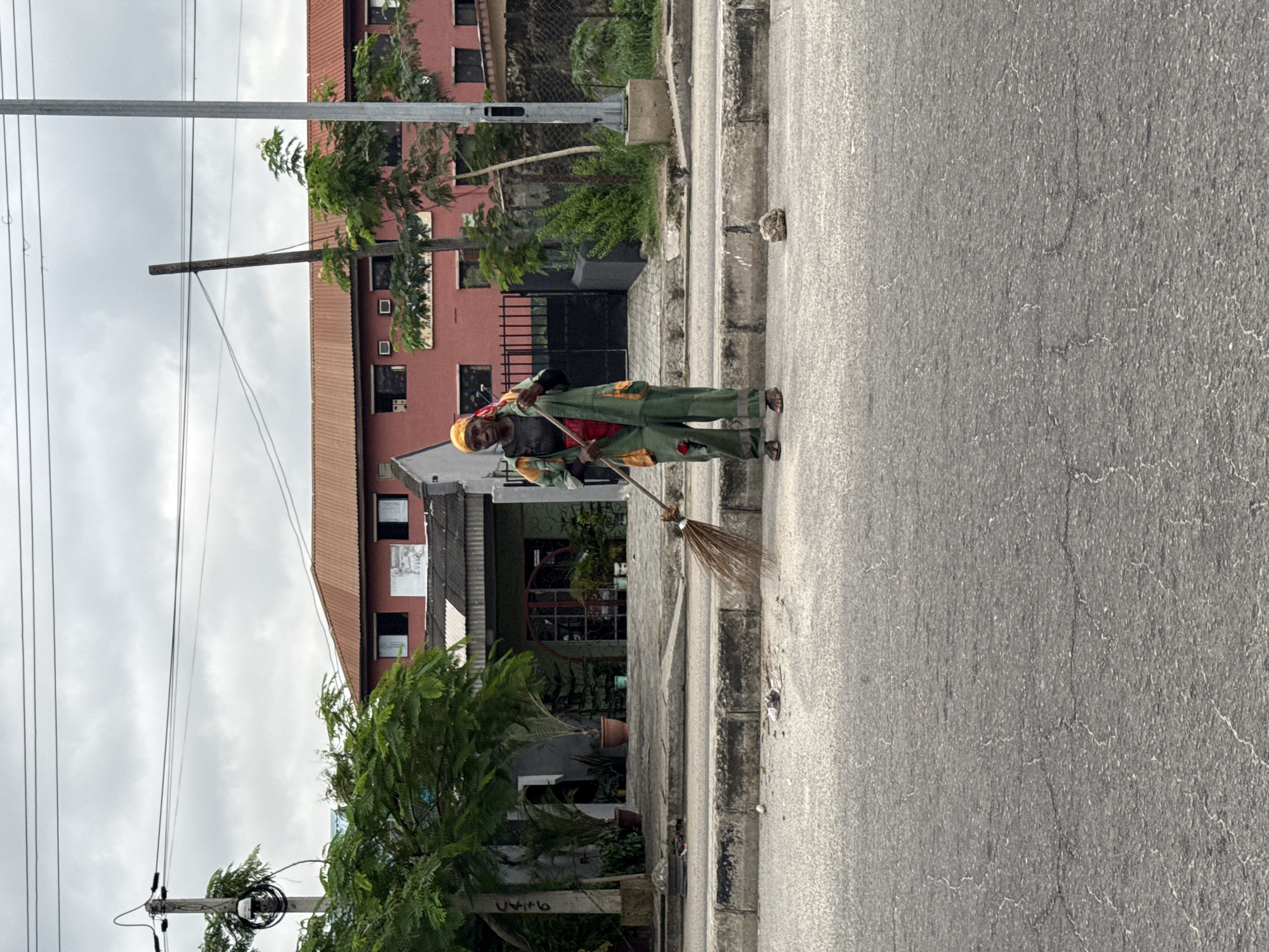 An individual, likely a street sweeper, is depicted in the process of sweeping a paved road in an urban setting identified as Ado, Nigeria. The person is dressed in a distinctive green and yellow uniform or jumpsuit, with a red shirt visible underneath and a yellow head covering. They are wearing sandals and are holding a traditional broom made of natural fibers, positioned near the curb as if actively sweeping debris.
The scene takes place outdoors during the daytime, under an overcast or partly cloudy sky, suggesting bright but diffused sunlight. To the right of the person, there's a concrete curb and a sidewalk. Beyond the sidewalk, various buildings and lush green trees form the background. A prominent multi-story building with reddish-brown walls and a brown corrugated roof is visible, along with other structures.
The environment is characterized by overhead utility poles and numerous power lines stretching across the frame, a common sight in urban areas of the region. The road surface is asphalt, displaying some minor cracks. The overall atmosphere is one of daily routine and urban maintenance. No clearly legible text or signage is visible in the image.