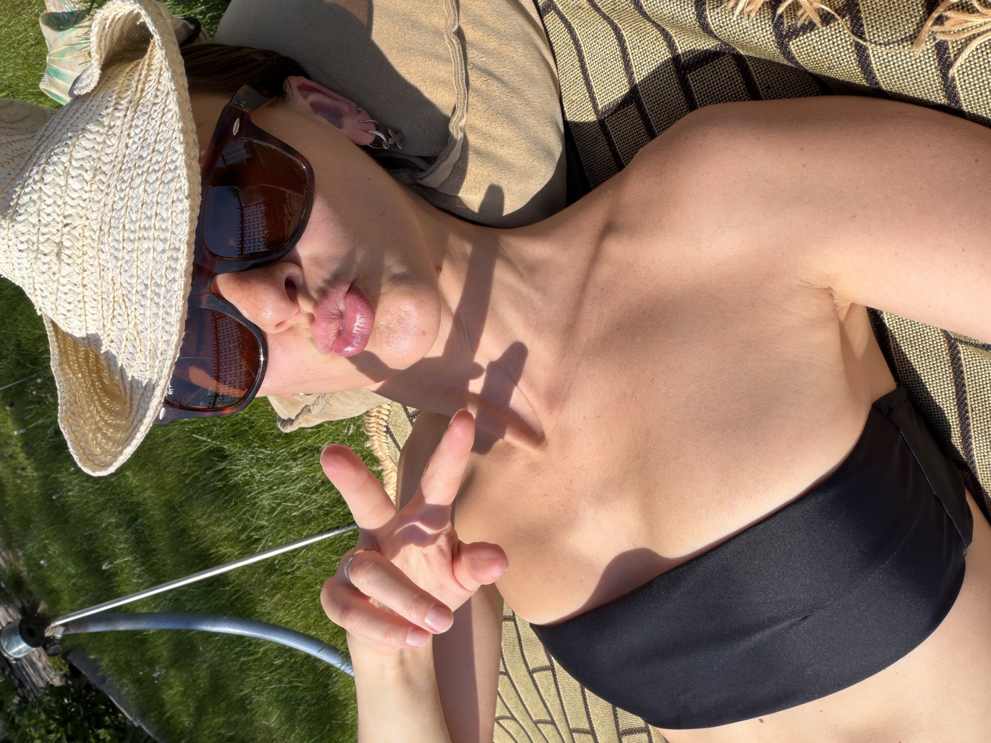 A woman lies on a striped beach mat, wearing a black bandeau bikini top, sunglasses, and a straw hat. She is positioned outdoors, likely on grass. She makes a peace sign with her hand. The location is not discernible as Kamyshin, Russia from this image alone.