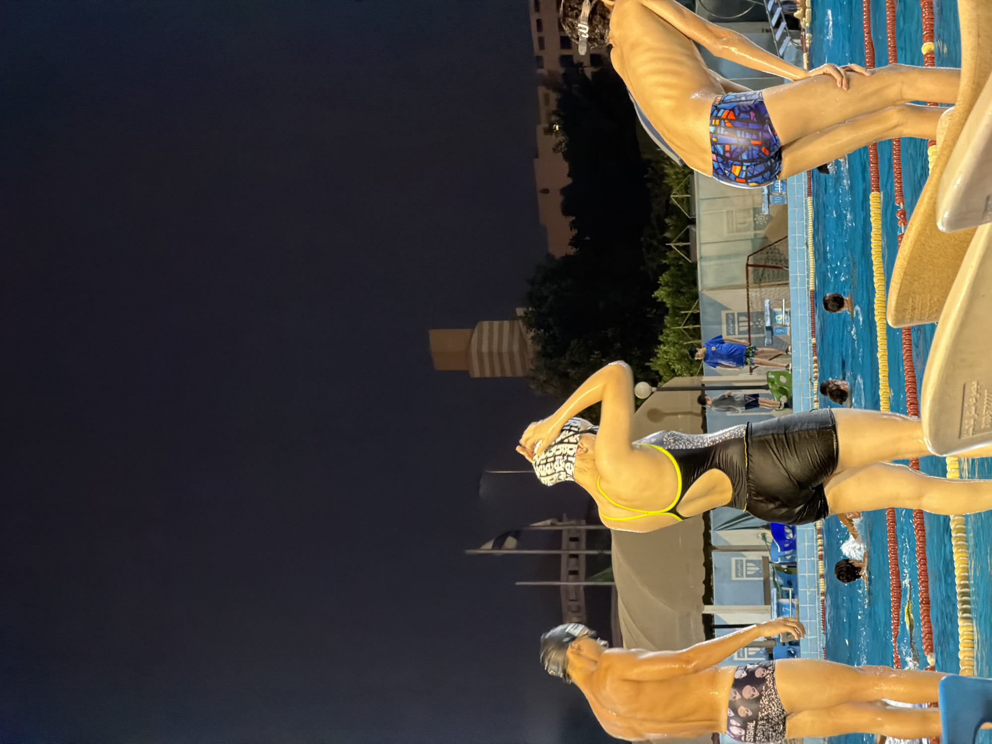 At nighttime, at a public swimming pool, a group of people are engaged in activities related to swimming. The scene is located in the city of 6th of October, Egypt, as evidenced by the architectural style of the buildings in the background and the presence of Egyptian flags. In the foreground, three individuals are prominently featured. On the left, a young man, wet and glistening, wears swim trunks with a skull pattern. Next to him, a woman is adjusting her swim cap, dressed in a black one-piece swimsuit with yellow accents. To the right, another young man, also wet, is wearing colorful swim trunks. In the background, other swimmers are visible in the pool. The pool is marked with lane lines and floats. The surrounding environment includes trees, buildings, and lighting fixtures, all illuminated against the dark night sky.