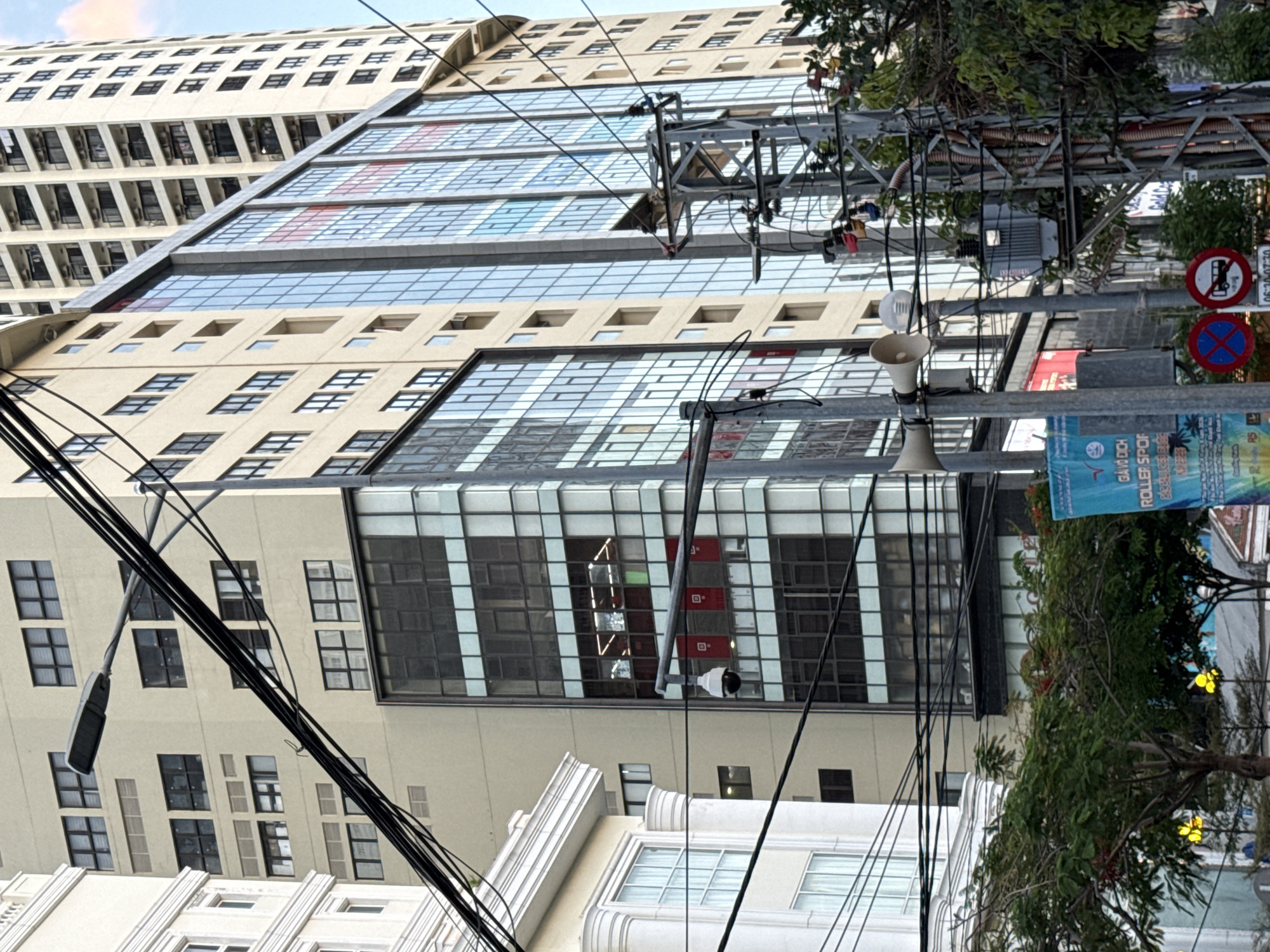 The image depicts a street scene in Nha Trang, Vietnam, dominated by tall buildings and urban infrastructure. In the foreground, a network of thick black electrical wires crisscrosses the frame, suspended from metal poles. A security camera is visible on a pole, facing left. The buildings in the background vary in architectural style. To the left, a cream-colored building with classical architectural elements, featuring arched windows and decorative molding, stands prominently. Adjacent to it is a modern building with a beige facade and numerous rectangular windows. Further to the right, a tall, modern building with a glass facade, reflecting the sky and surrounding structures, rises high. Red and white rectangular banners are visible behind the glass on several floors. At the base of the modern buildings, trees with green foliage and visible red flowers add a touch of nature to the urban environment. Street signs and what appear to be speakers are also mounted on poles. The overall impression is a busy, commercial urban area.