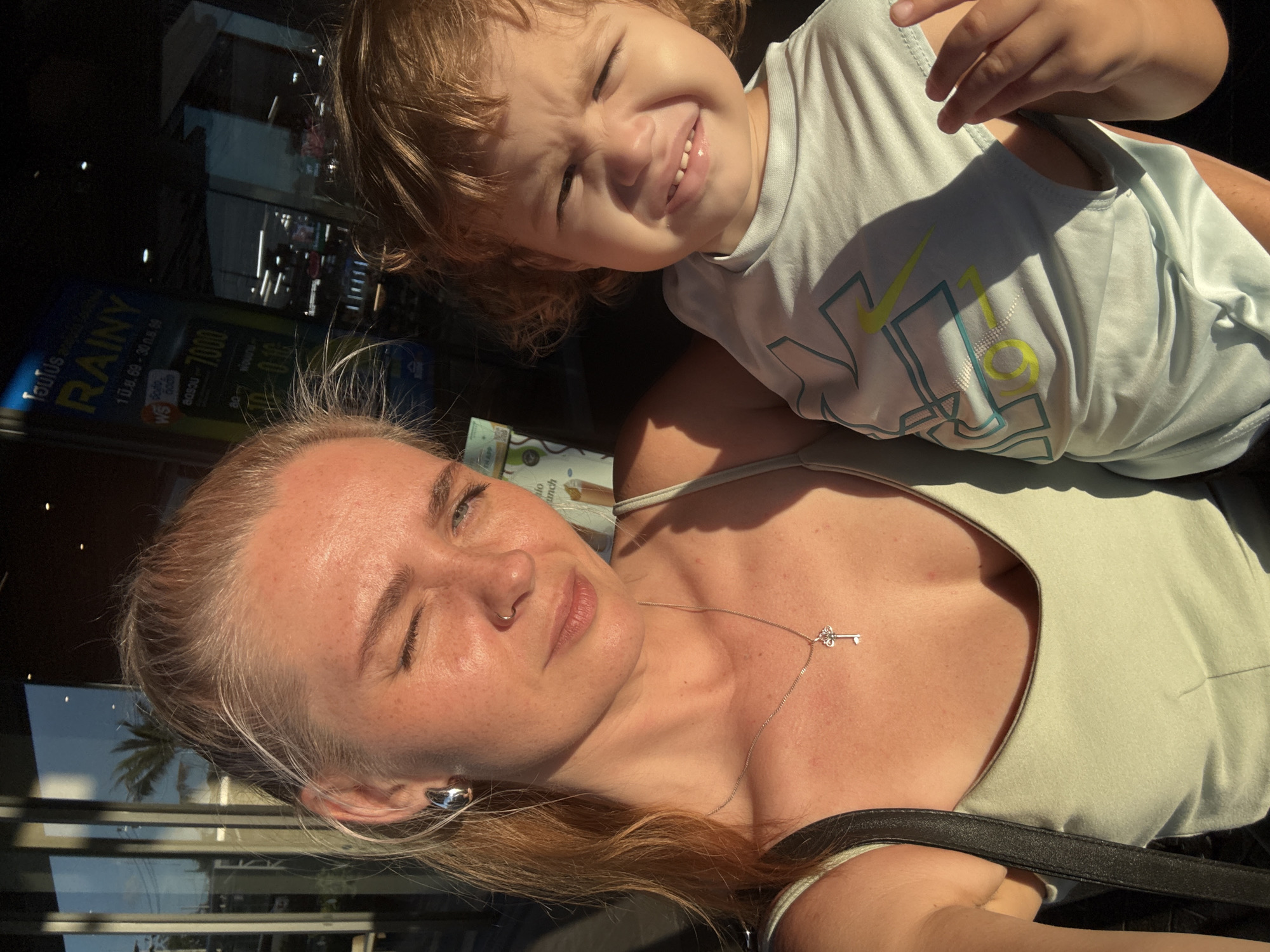 In Choeng Thale, Thailand, a woman with blonde hair pulled back in a ponytail is holding a young child. The woman is wearing a light green tank top and a silver necklace with a key pendant. She is also wearing a silver hoop earring. The child is wearing a light blue t-shirt with a graphic design. Both individuals are looking towards the camera, and the woman is winking. Sunlight is casting shadows on their faces. In the background, there is a building with reflective windows, and a sign with Thai writing and the word "RAINY" is visible. The scene appears to be outdoors, possibly near a commercial establishment.