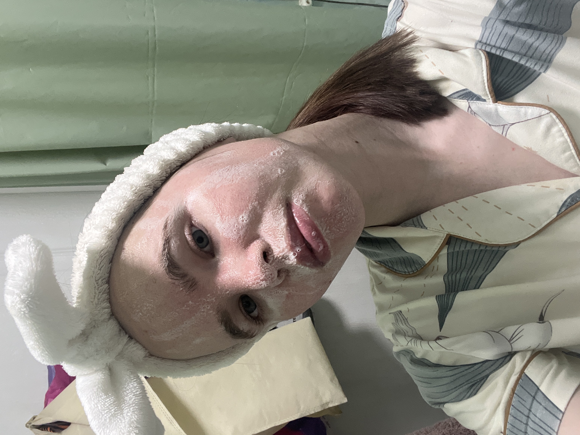 An eye-level, tilted portrait captures a person engaged in a skincare routine indoors, likely in Chaikovsky, Russia. The subject, who appears to be female, has fair skin and dark brown hair, which is pulled back. She is wearing a white, fluffy terry cloth headband with bunny ears, securing her hair away from her face. Her face is covered with a white, foamy substance, indicative of a face mask or cleanser, with small bubbles visible on her cheeks, nose, and lips. Her eyes are open and gaze directly towards the viewer, showing a neutral expression.
She is dressed in a light-colored pajama top with a cream background and a pattern of grey-blue, leaf-like or abstract shapes, accented with brown piping. Her neck and a portion of her shoulder are visible. The background features a muted green, possibly a curtain or blind, suggesting a window or an interior wall. Below her head, a beige or cream-colored fabric item is partially visible. The overall setting implies a private, relaxed moment, possibly in a bedroom or bathroom. There is no visible text in the image.