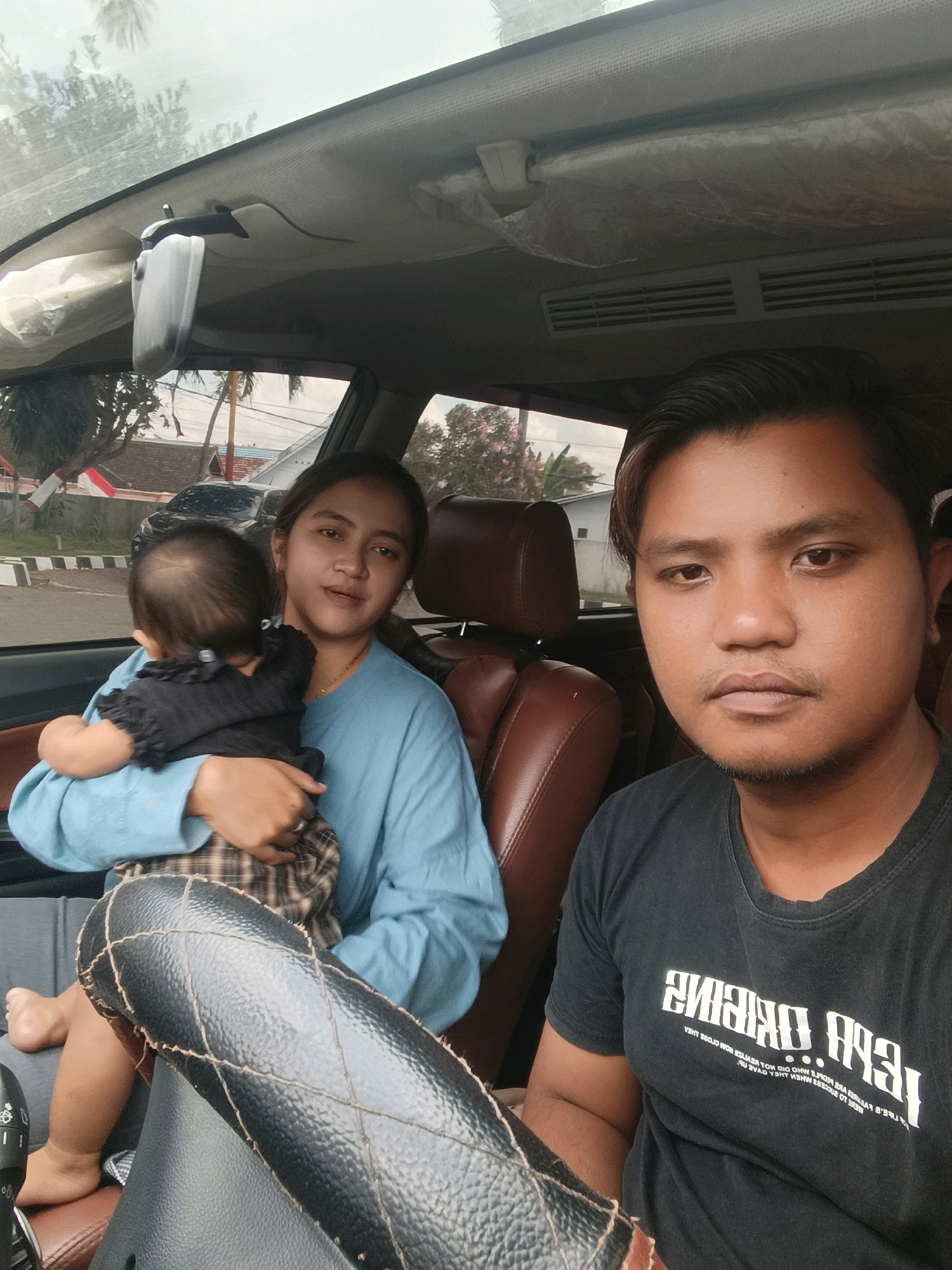 In Tempeh Lor, Indonesia, a man and a woman are seated in the front seats of a vehicle. The woman, on the left, holds a baby, facing away from the camera. The man, on the right, faces forward, looking towards the camera. The interior of the car is visible, including the dashboard, steering wheel, and seats. Outside the vehicle, trees, houses, and a road are discernible, suggesting a semi-urban or suburban setting. The lighting indicates it is daytime.