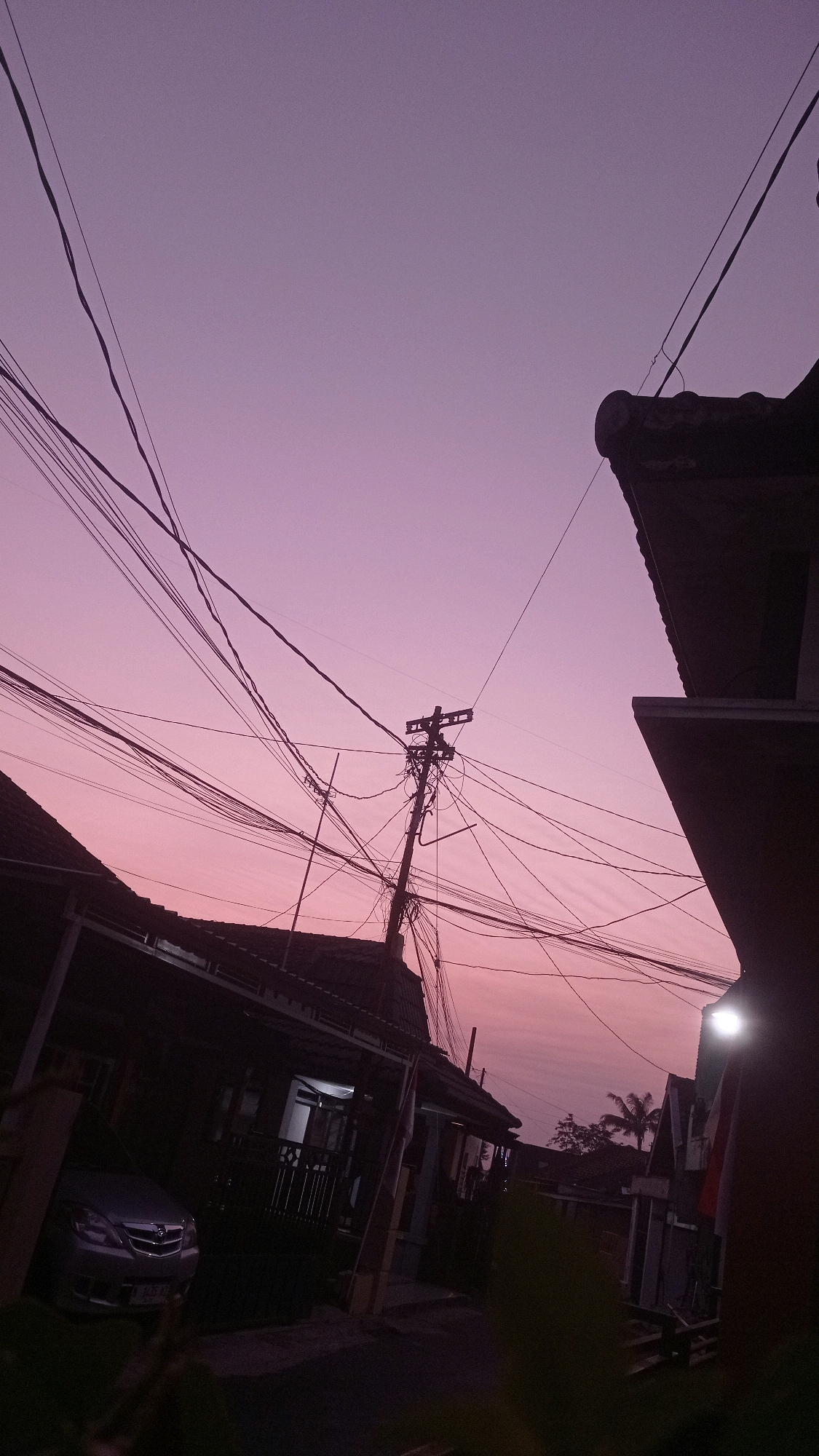 The image depicts a street scene at dusk in Mulyoagung, Indonesia. A silver car is parked in front of a house. Overhead, a complex network of electrical wires crisscrosses the sky, suspended from a utility pole. The sky is a soft gradient of purple and pink, characteristic of twilight. Two silhouetted houses, one on the left and one on the right, frame the street. The house on the right has a bright outdoor light illuminating a portion of its facade. The scene is quiet, with no people visible.