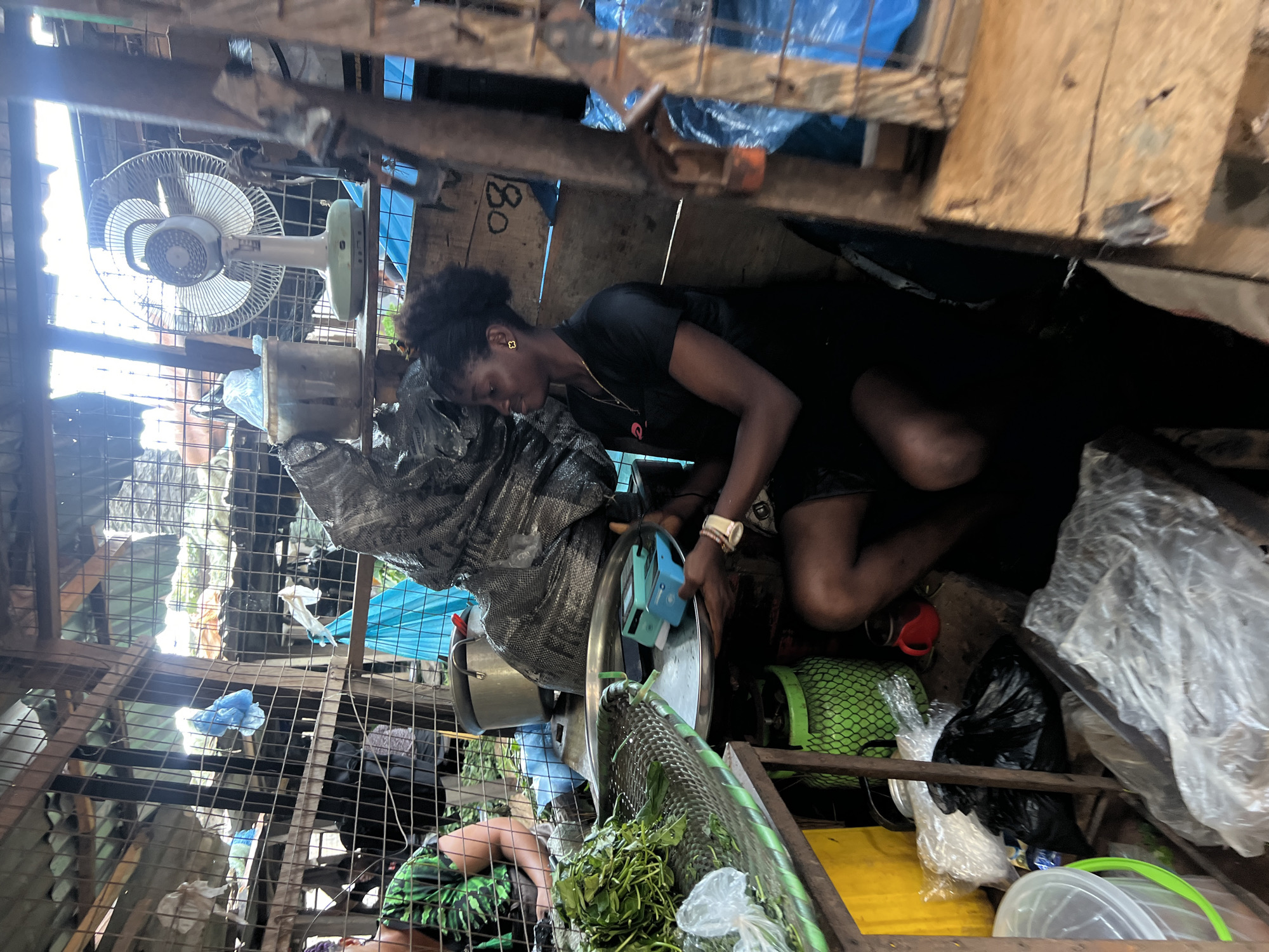 In Nkpor, Nigeria, a Black woman with dark, curly hair pulled back sits in a crowded marketplace stall. She is wearing a black t-shirt and dark shorts, and her legs are bent as she crouches. In her hands, she holds a large silver tray containing two small, teal electronic devices. To her left, a pile of green leafy vegetables is displayed on a green-rimmed surface. Behind her, a large, grey, crinkled bag dominates the background. The stall is constructed with rustic wooden beams and corrugated metal roofing, with various items including plastic bags, a red mug, and a green propane tank scattered around. A white electric fan is visible on a shelf in the upper right corner. The scene depicts a typical market setting, likely a vendor preparing for or engaged in business.