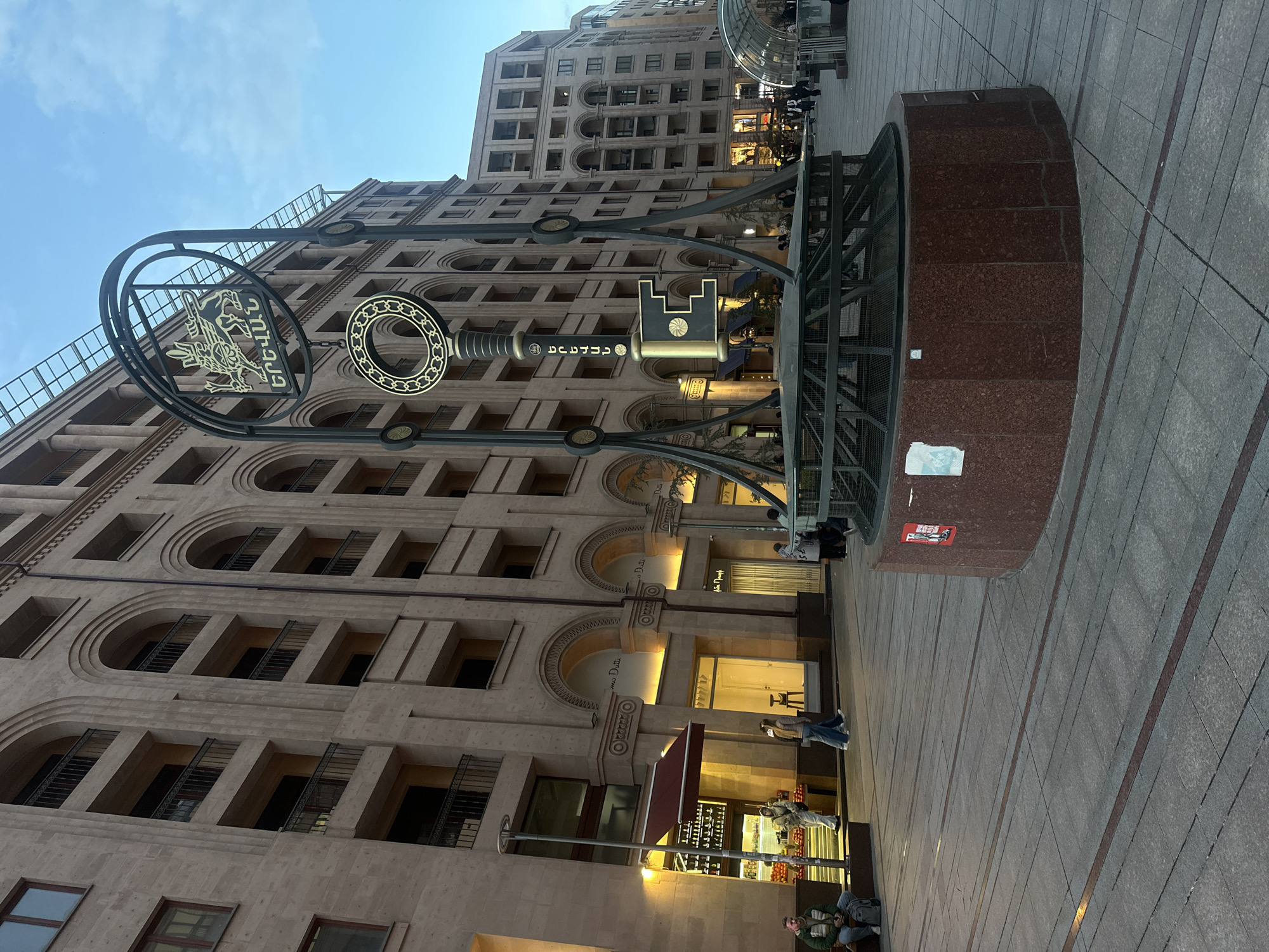 The image shows a street scene in Yerevan, Armenia, dominated by a large, ornate metal sculpture in the shape of a key. The sculpture features decorative elements and text in Armenian script. The background features a multi-story building with arched windows and a facade constructed from light-colored stone or concrete.
People are present on the street, with some visible near storefronts. The street is paved with rectangular stones. The sky is visible in the upper portion of the frame. The scene appears to be lit by both natural and artificial light sources.