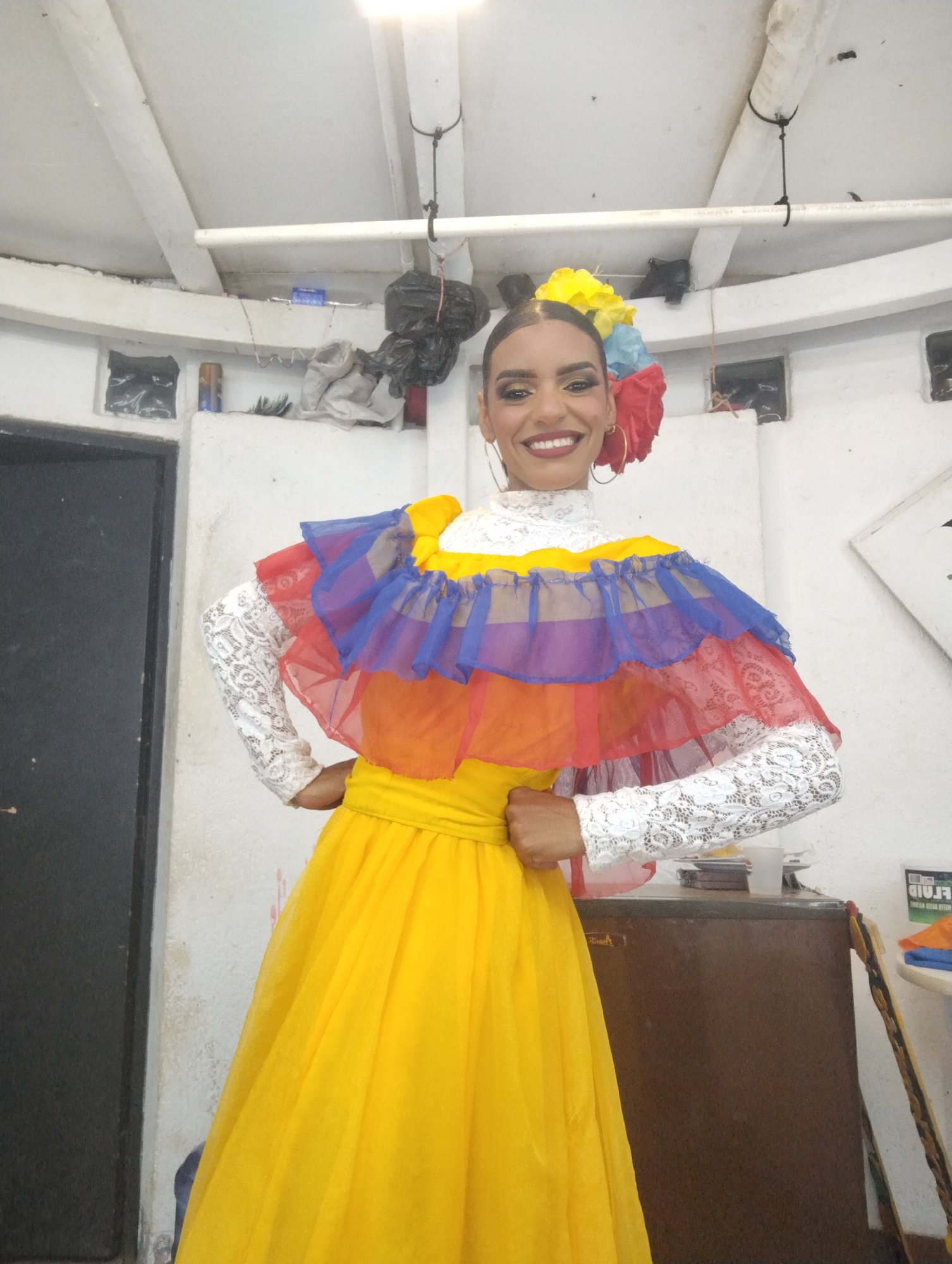A woman stands smiling in an interior space. She is wearing a traditional dress featuring a yellow skirt and a top with lace sleeves and a ruffled collar in the colors of the Venezuelan flag: yellow, blue, and red. Her hair is styled up with colorful flowers, and she wears large hoop earrings. The background consists of a white-painted room with a dark door to the left, various items stored on a shelf overhead, and a brown cabinet to the right. The environment suggests a backstage or storage area. The location is likely in Porlamar, Venezuela, based on the traditional dress and inferred cultural context.