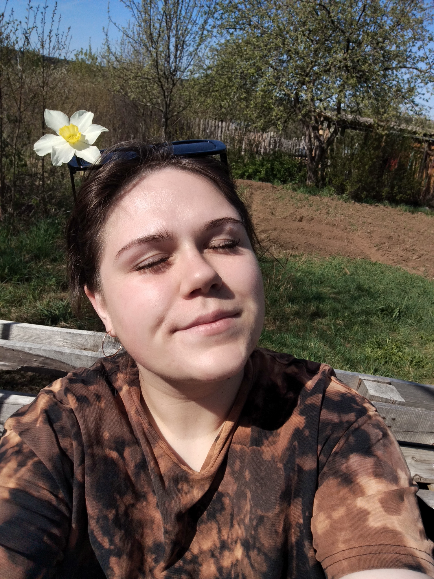 A person is photographed outdoors in Chaikovsky, Russia. The person has fair skin, brown hair, and wears a brown and black tie-dye t-shirt. Their eyes are closed. A white flower is placed on top of their head, near a pair of sunglasses. The background includes trees, a grass field, and what appears to be a cultivated garden. The person is sitting on a wooden structure.