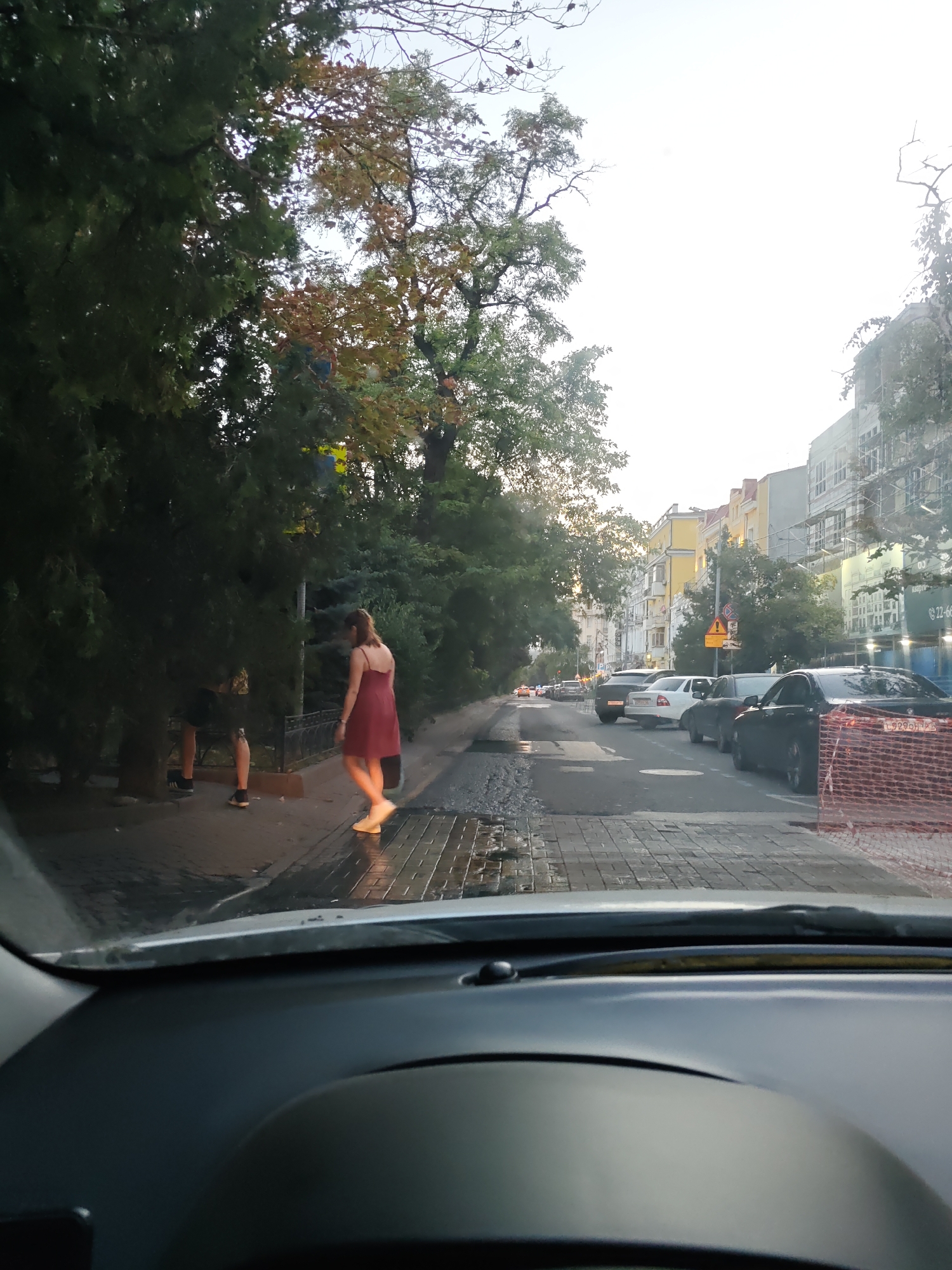 The image shows a street scene in Rostov-on-Don, Russia. The perspective is from inside a car, looking out through the windshield. In the foreground, the dashboard of the car is visible. The street is paved with cobblestones and appears to be wet, with puddles of water. On the sidewalk, there are trees with green and autumn-colored leaves. Two individuals are partially visible on the sidewalk, obscured by trees. One person, a woman, is walking away from the camera, wearing a red dress and white shoes. Another person, a man, is standing partially behind trees, wearing shorts and dark shoes.
The street is lined with parked cars, including a black BMW in the foreground. Buildings line the opposite side of the street, some appearing to be under construction with scaffolding. The sky is bright, suggesting it is daytime. The overall atmosphere appears to be that of a residential or mixed-use urban area. The license plate of the black BMW appears to be from Russia, possibly with a regional code for Rostov Oblast.